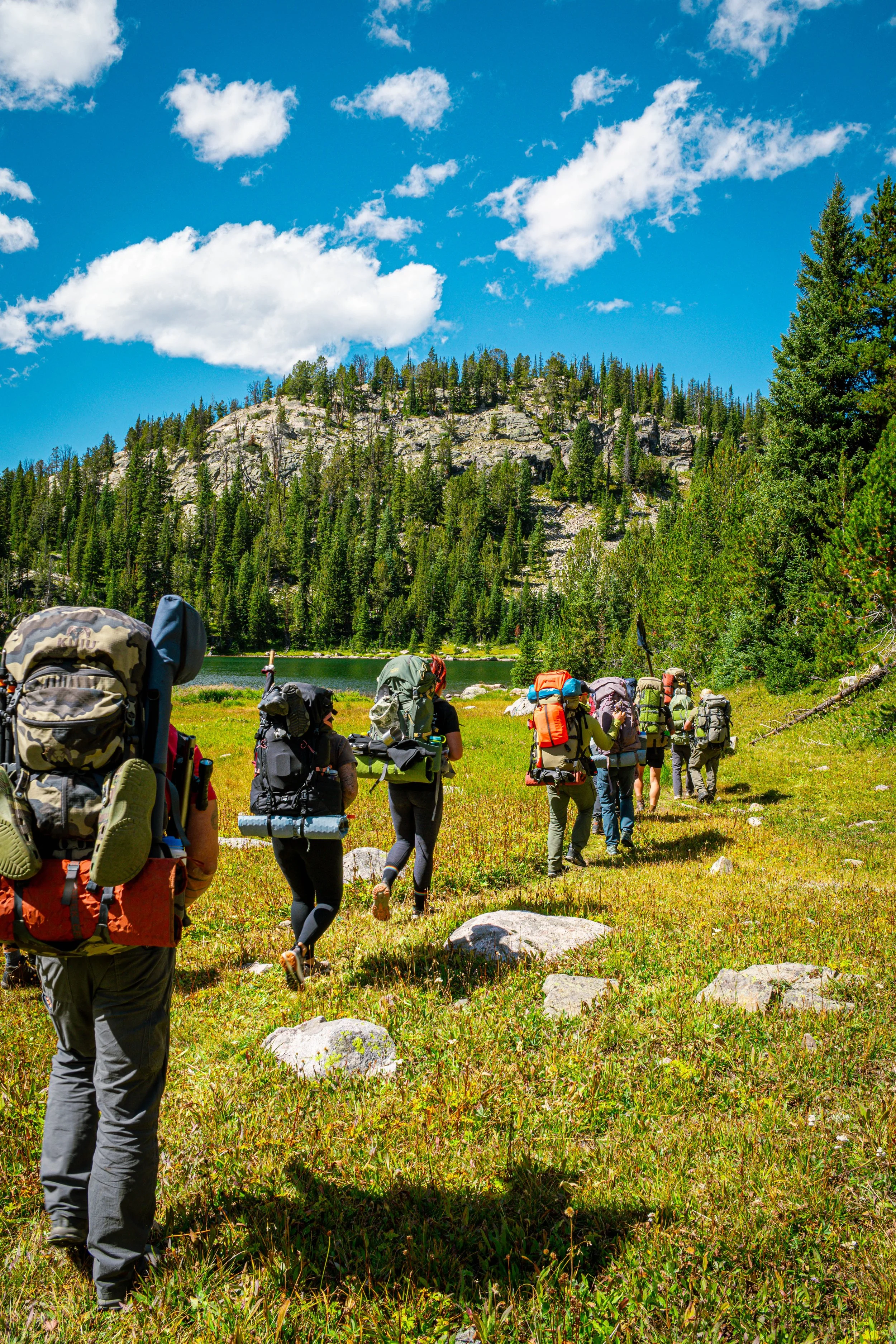 Group of veteran hikers with backpacks walking along a grassy trail beside a lake, with a rocky hill and pine trees in the background under a partly cloudy sky.