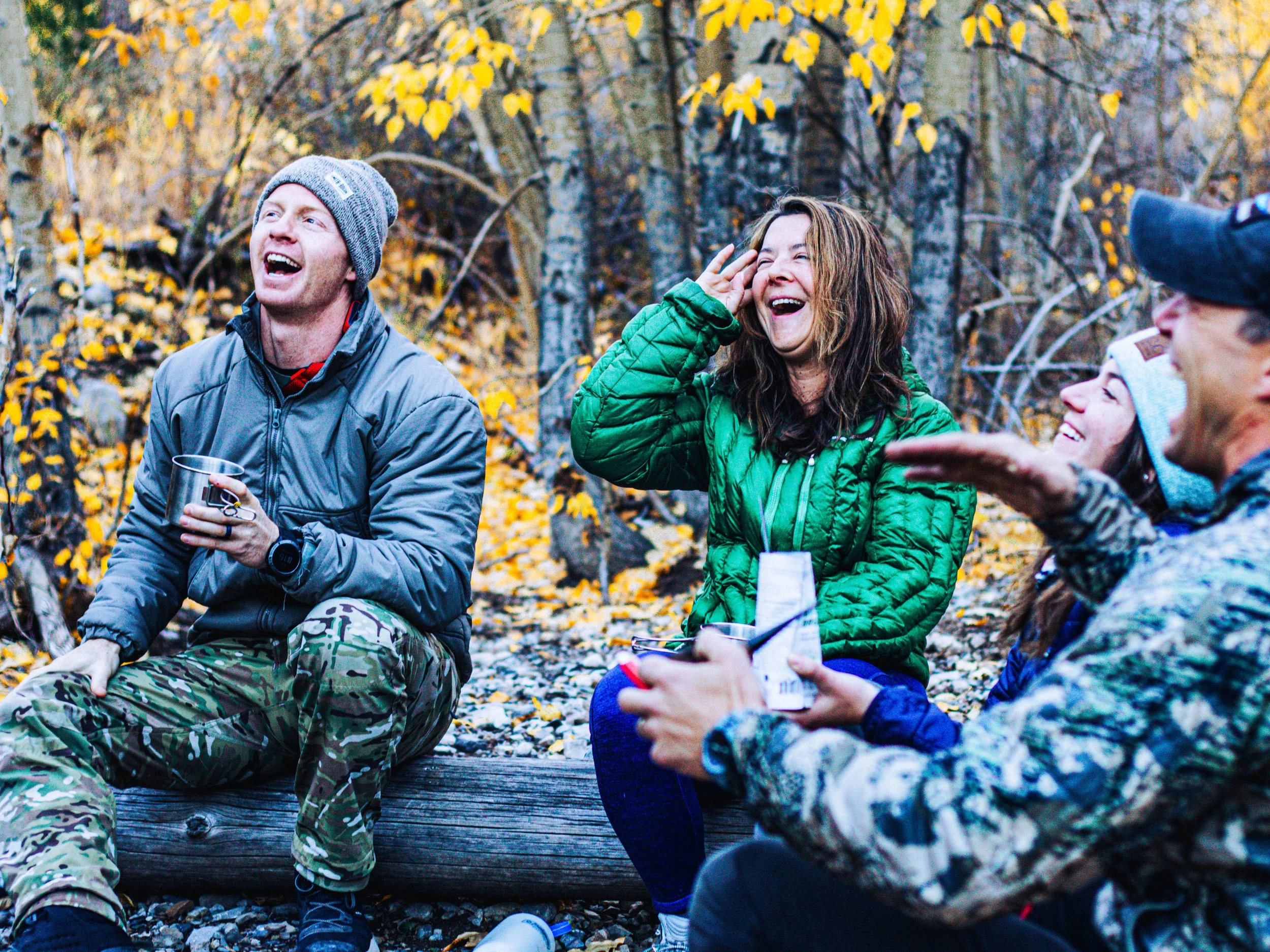 Group of veterans sitting outdoors in a wooded area during fall, laughing and talking, wearing jackets and hats.