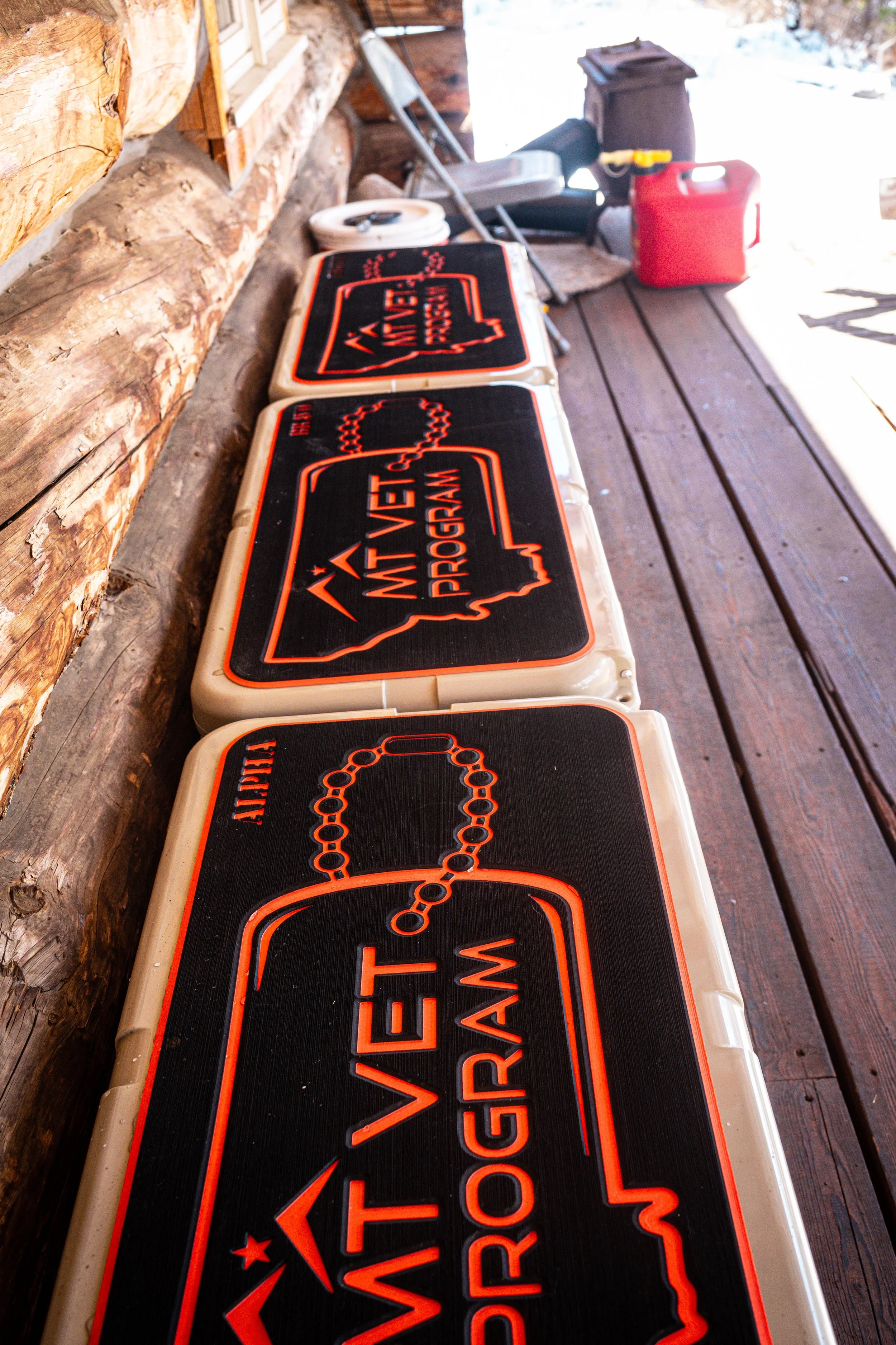 Close-up of multiple warning signs for a motorcycle race, placed on a wooden porch, with a log wall on the left and a red gas can and other items on the right in the background.
