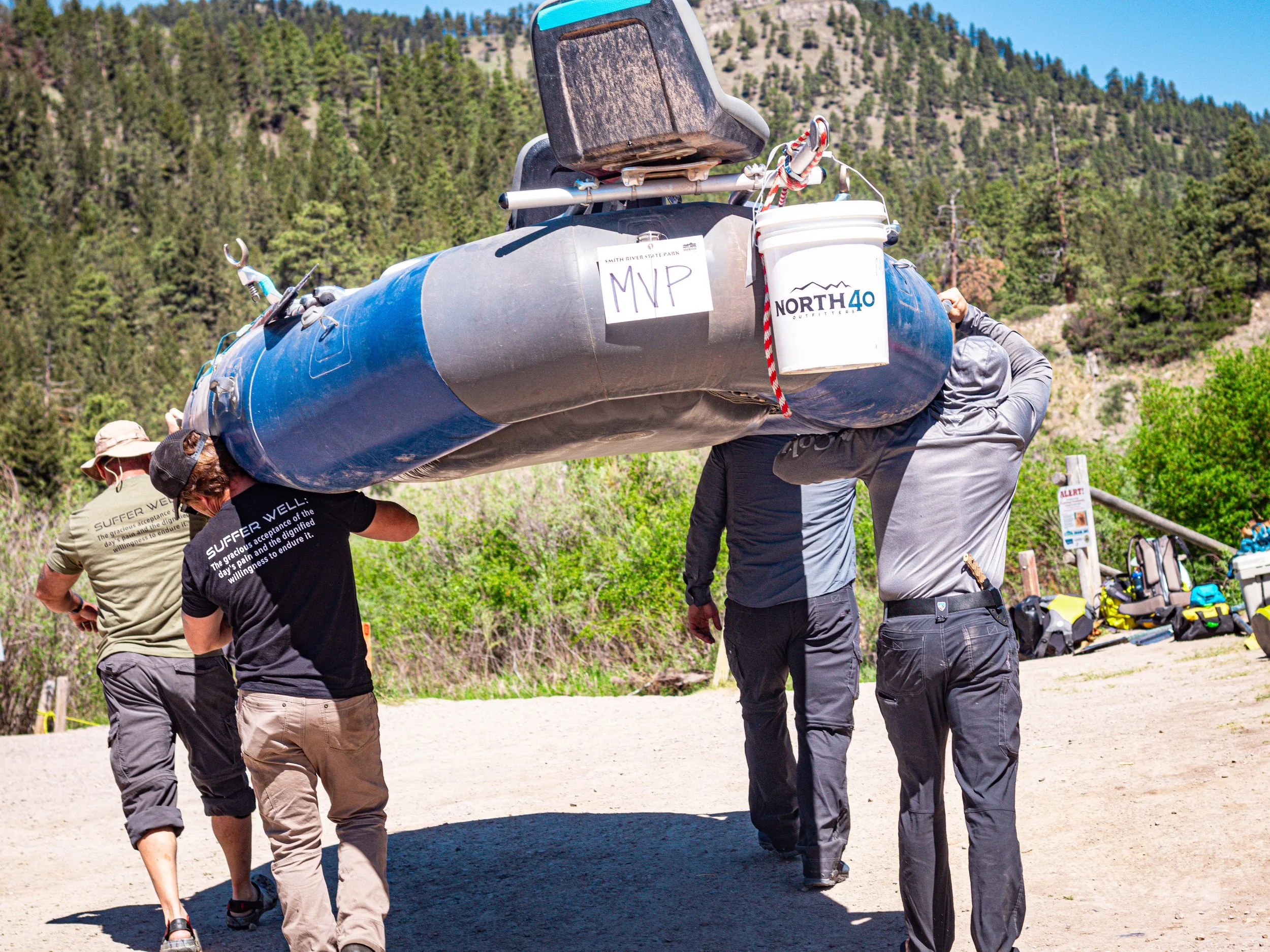 A group of people carrying a kayak on their shoulders outdoors in a mountainous area with green trees and a clear blue sky.