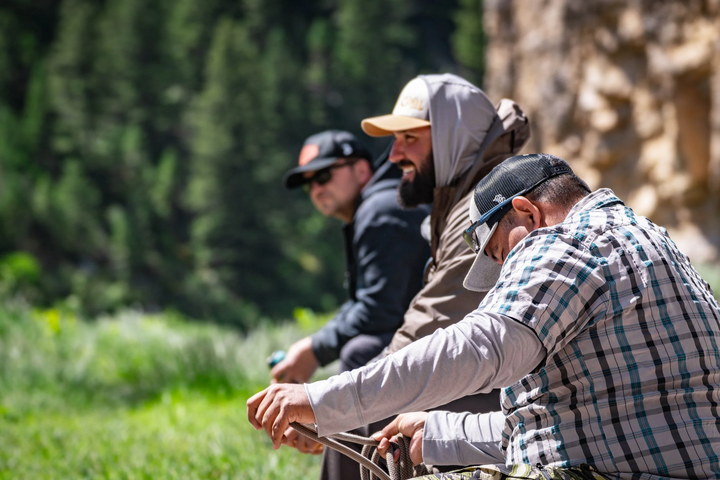 Three men sitting outside in a natural setting, smiling and engaging in conversation.