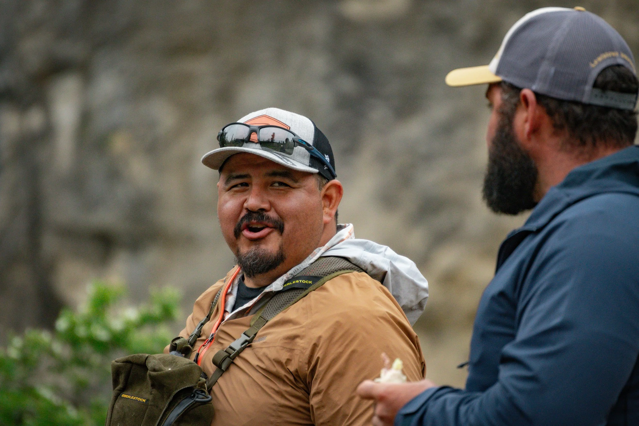 Two men engaged in conversation outdoors, one has a mustache and beard, wearing a beige jacket and a cap with sunglasses on top. The other man, visible only partially, has a beard, wears a blue jacket and a gray cap, and is holding a small object in his hand. The background is blurred with trees and natural landscape.