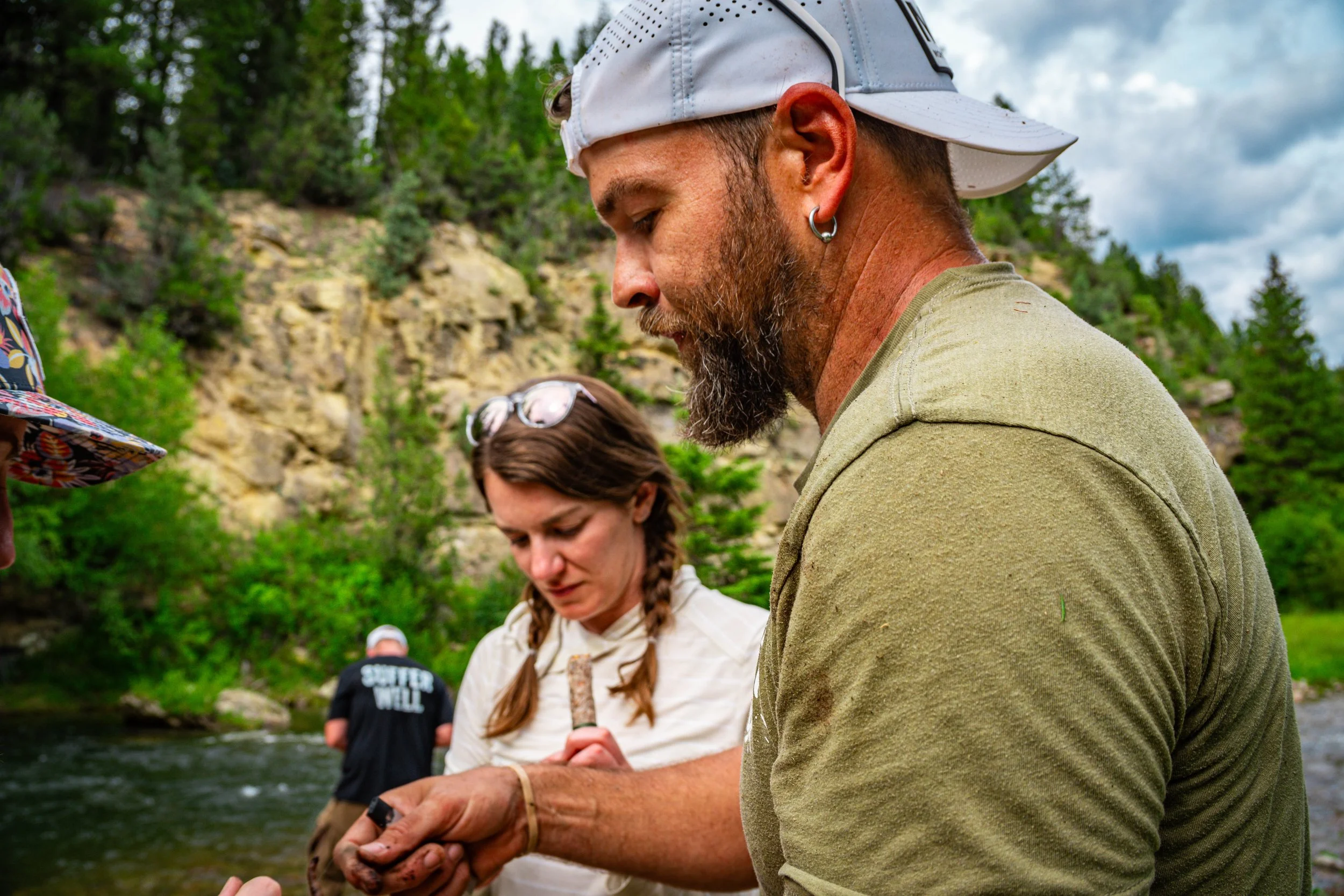 A group of people standing outdoors near a river, with a man in a beige shirt and white cap in the foreground, and women in the background, some holding small objects, all surrounded by green trees and rocky landscape.