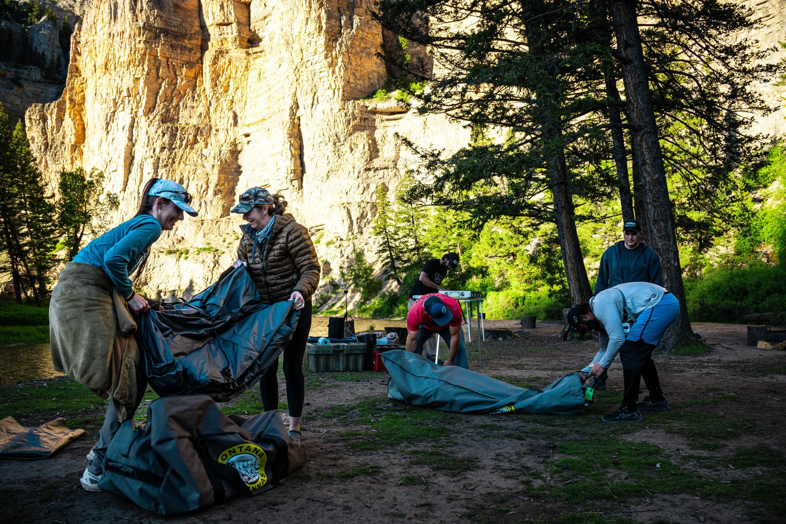 Group of people setting up camping gear near a river in a forested area with tall rock cliffs in the background.