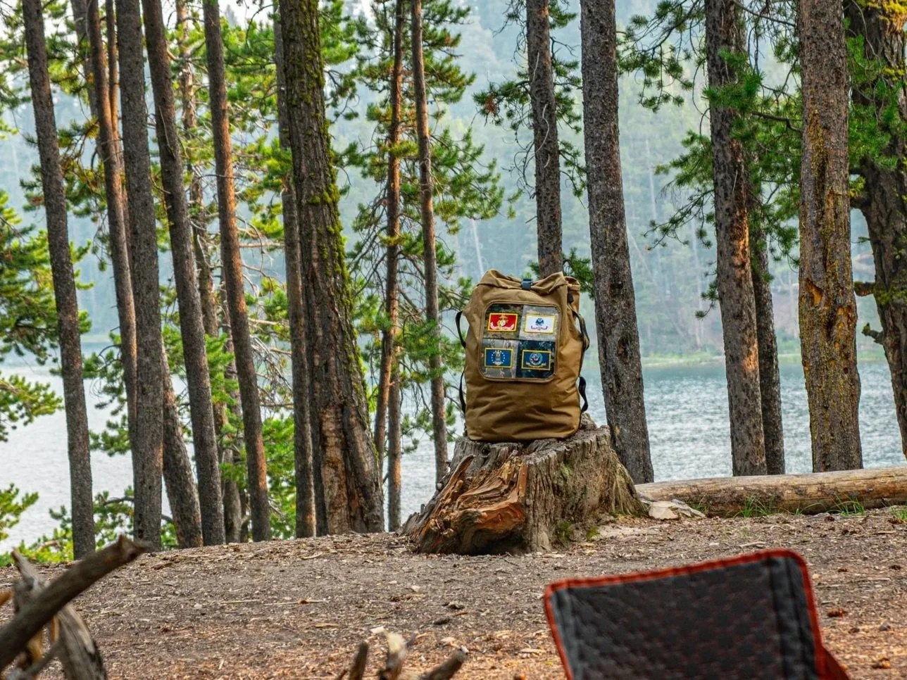 A tan backpack with patches sits on a tree stump in a forest near a lake, surrounded by tall pine trees.