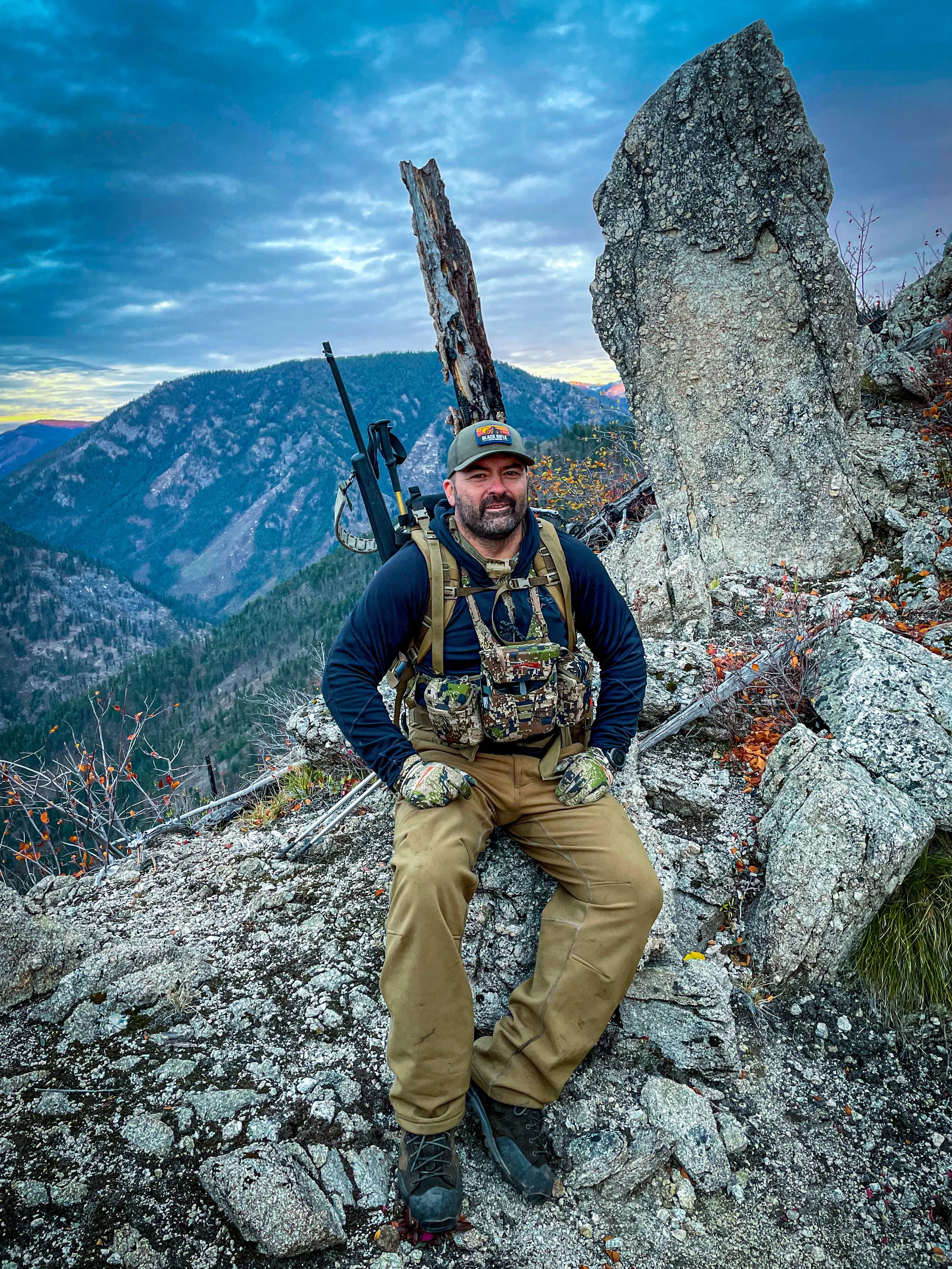 A man sits on a rocky mountain ledge, wearing outdoor gear, with mountains and a cloudy sky in the background.
