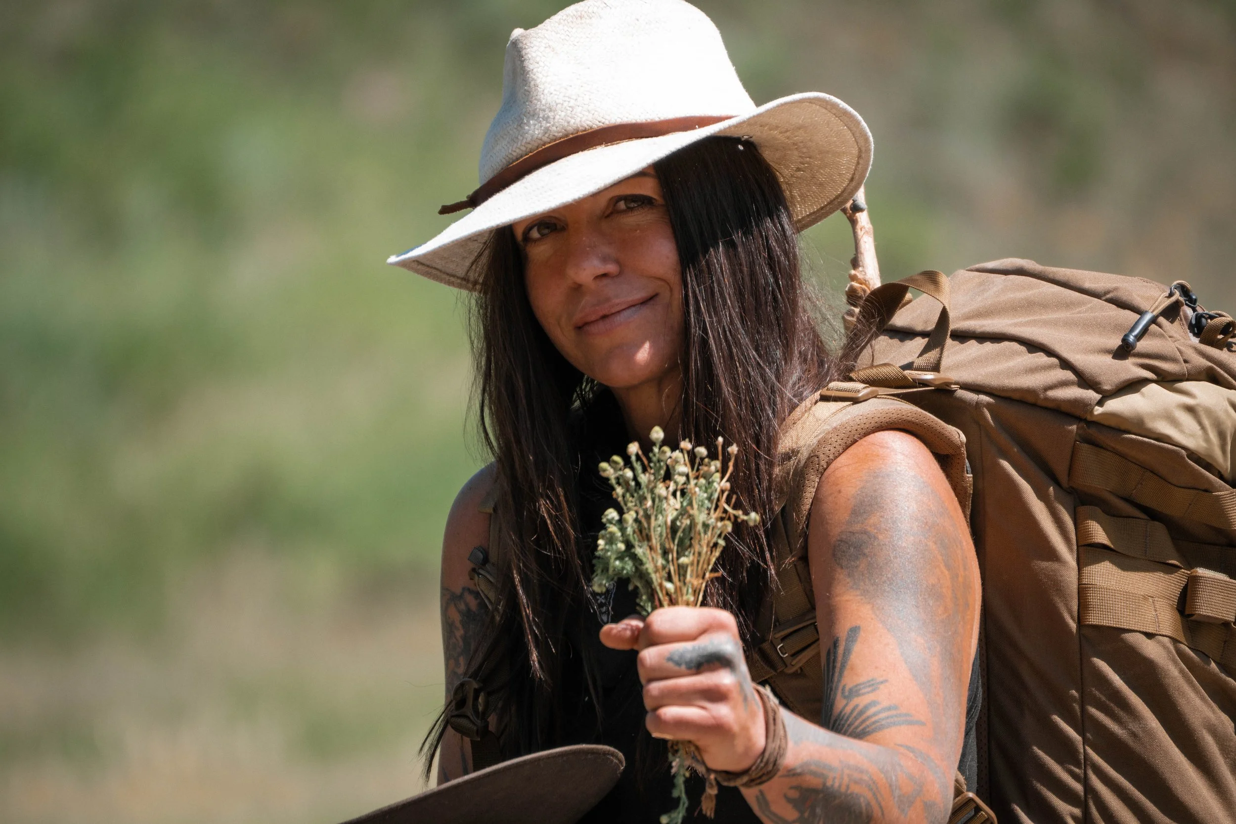Woman with long black hair wearing a beige wide-brim hat, carrying a backpack, holding a bunch of wildflowers, and wearing tattoos on her arms, outdoors in a green setting.