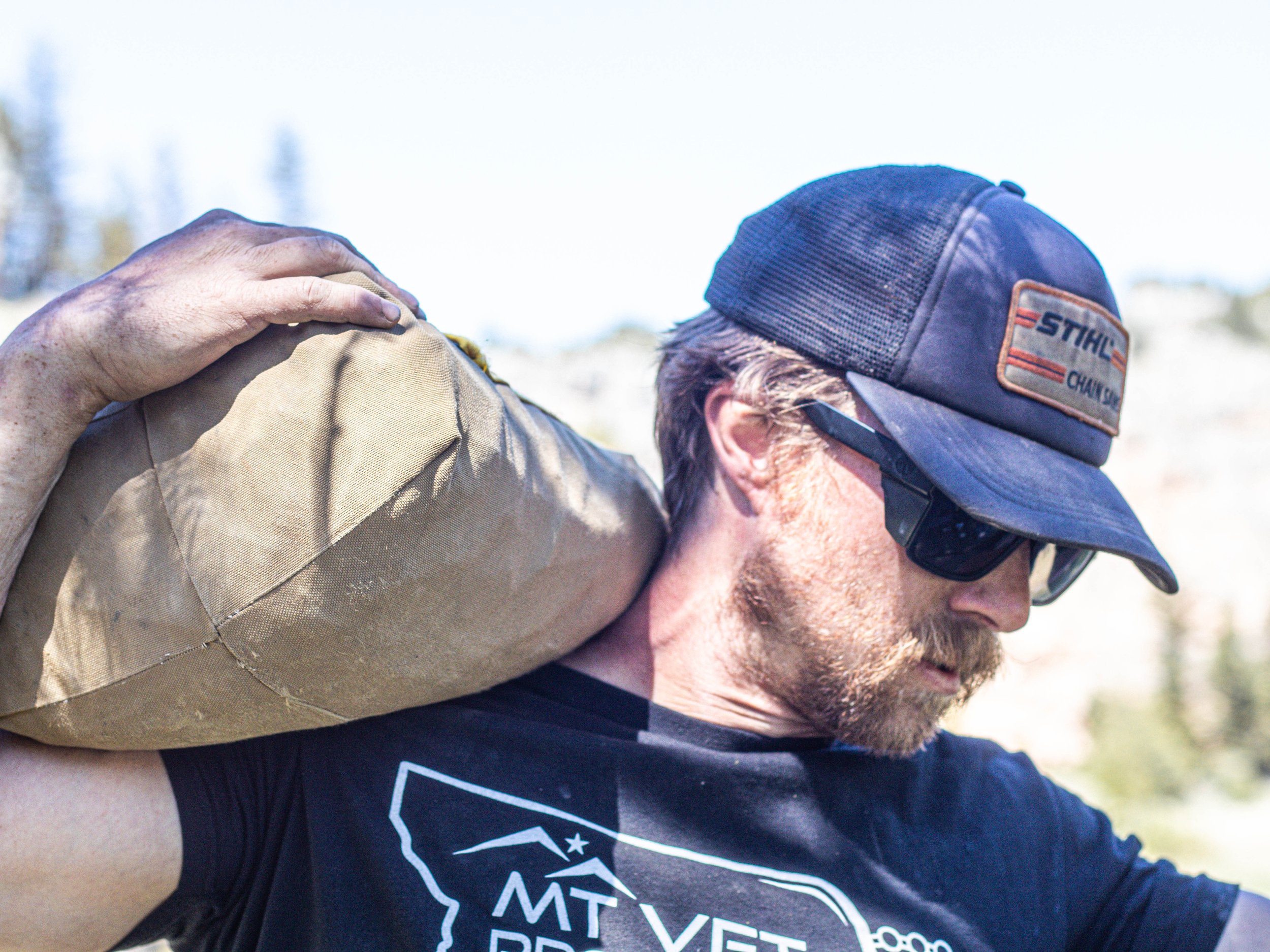 A person with a beard and sunglasses wearing a baseball cap with a 'STIHL' logo, assisting another individual on the shoulder in an outdoor setting.