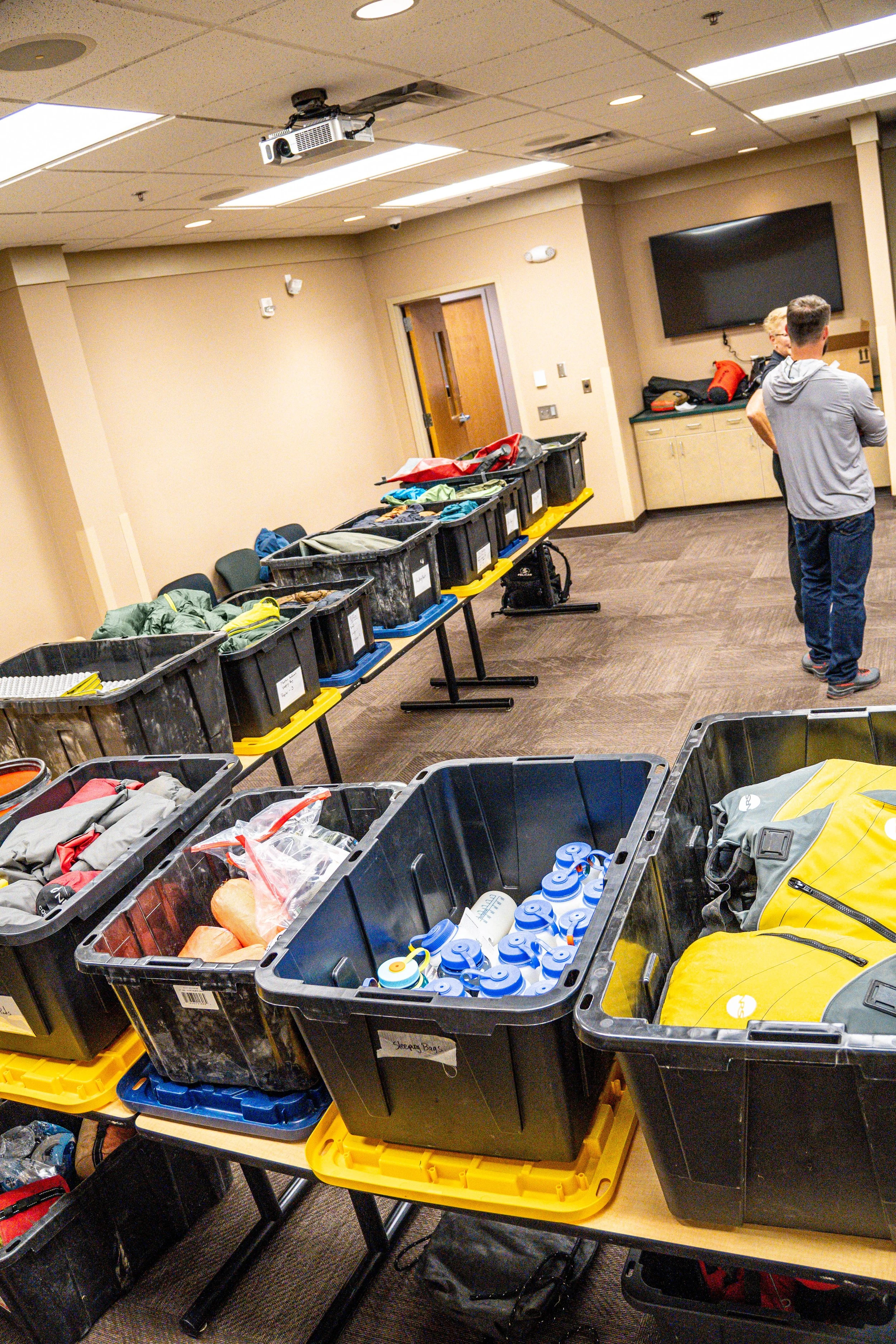 Table filled with black storage bins containing various outdoor clothing, gear, and water bottles in a conference room with beige walls and ceiling panels.