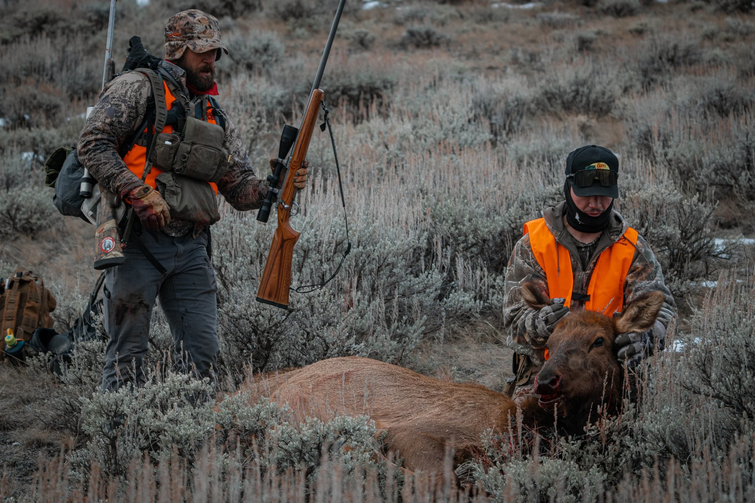 Two hunters in camouflage clothing and orange vests are in an open terrain with dry bushes. One is standing and holding a rifle, while the other is kneeling, petting a deer's head, which lies on the ground.