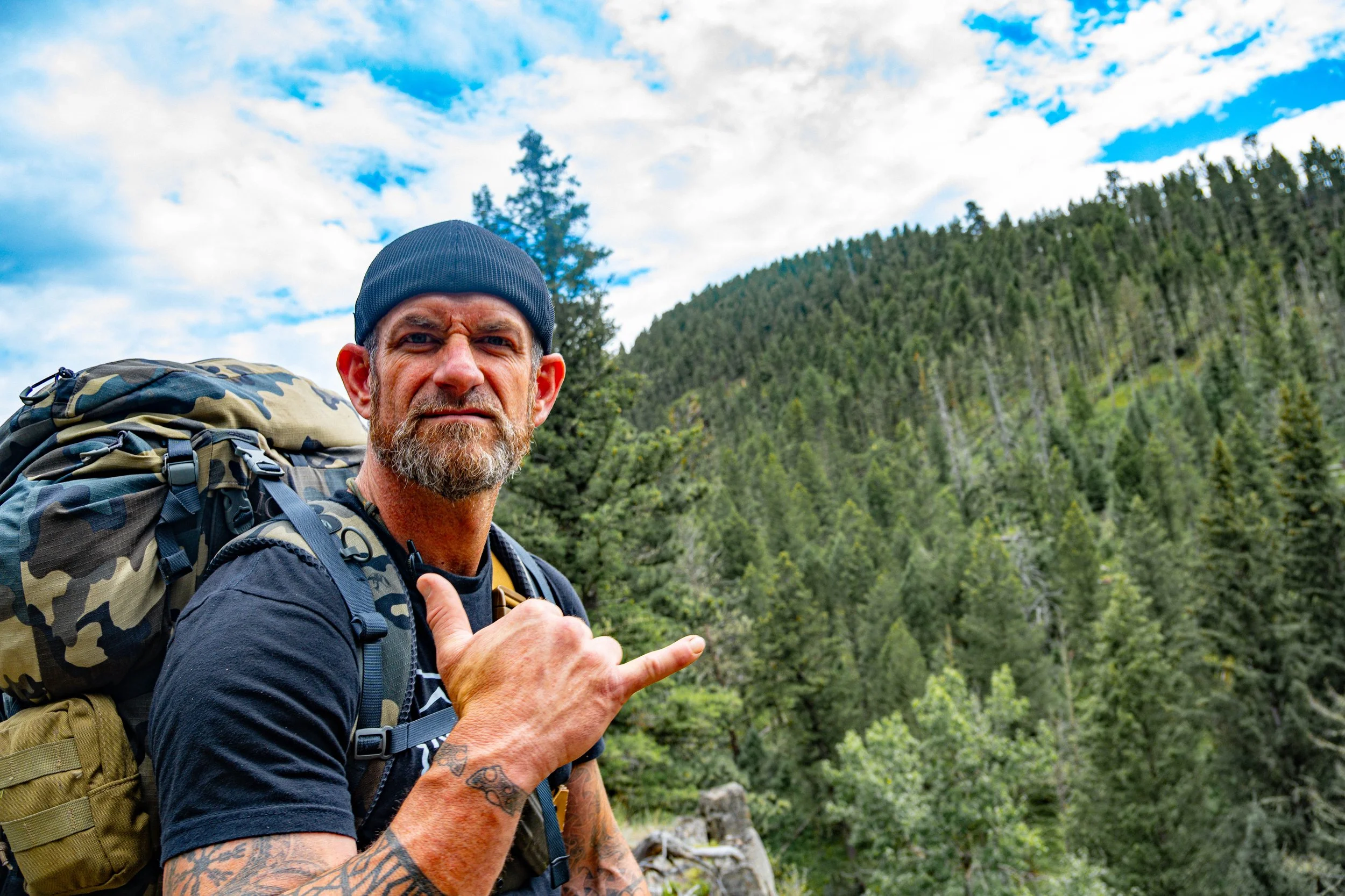 A man with a beard, wearing a black beanie and a black t-shirt, carrying a large camouflage backpack, giving a thumbs-up while standing outdoors in a forested area with blue sky and clouds.