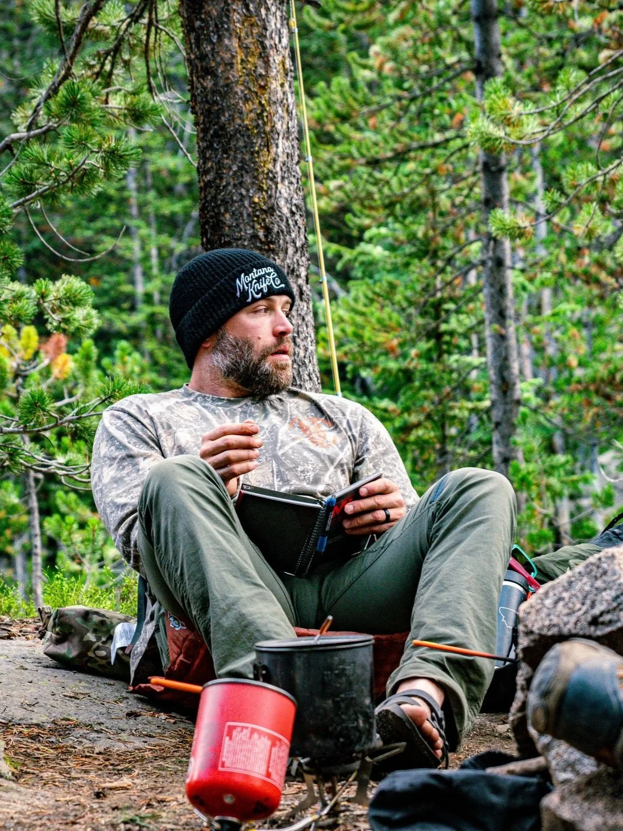 A man camping in a forest, sitting on the ground with a notebook in his lap, wearing a black beanie, camouflage shirt, and olive green pants. He's surrounded by camping gear, trees, and greenery.