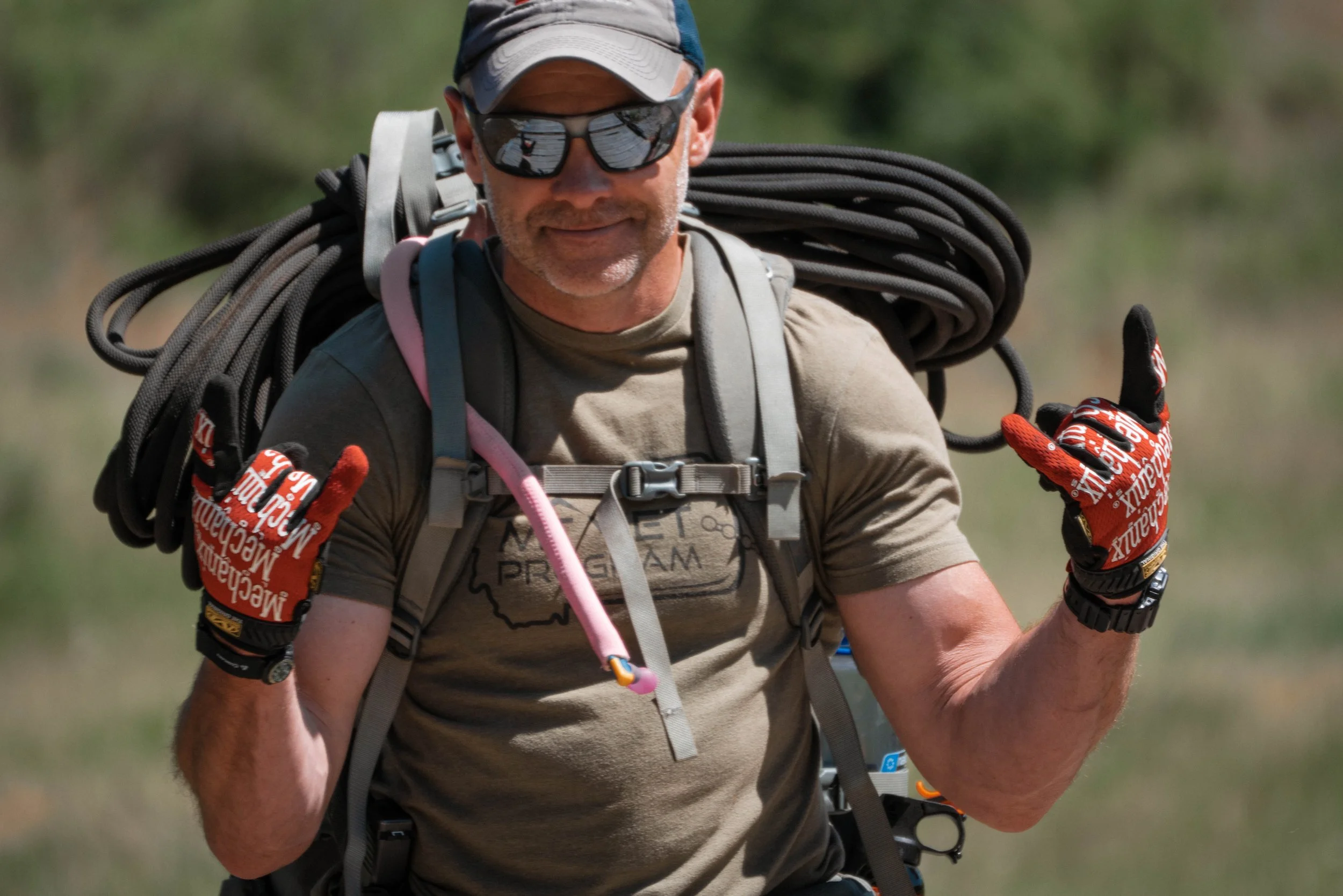 A man wearing sunglasses, a beige baseball cap, and beige T-shirt with a map of Wisconsin is carrying a large bundle of climbing ropes on his back while riding a bike outdoors.