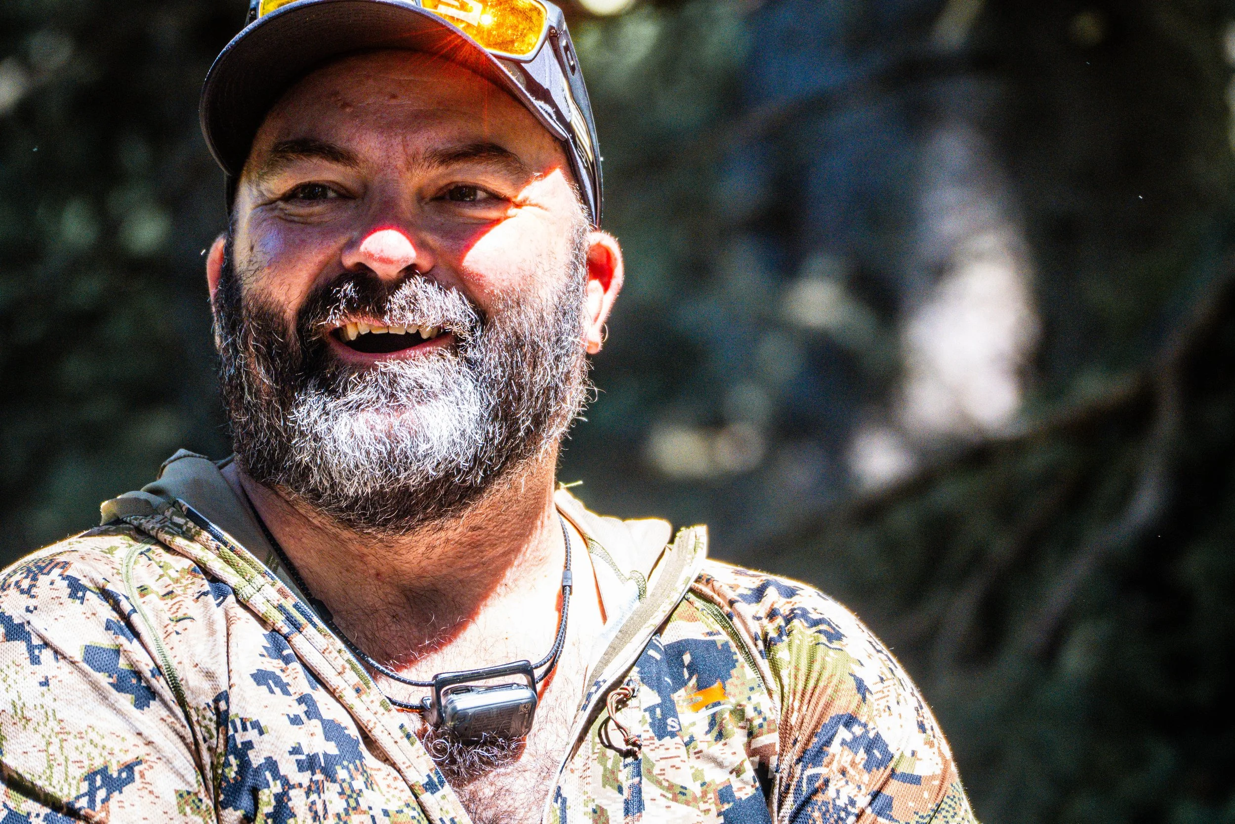 A smiling man with a beard and mustache, wearing a cap, sunglasses, a camouflage patterned shirt, and a GoPro camera hanging around his neck, outdoors with blurred trees in the background.