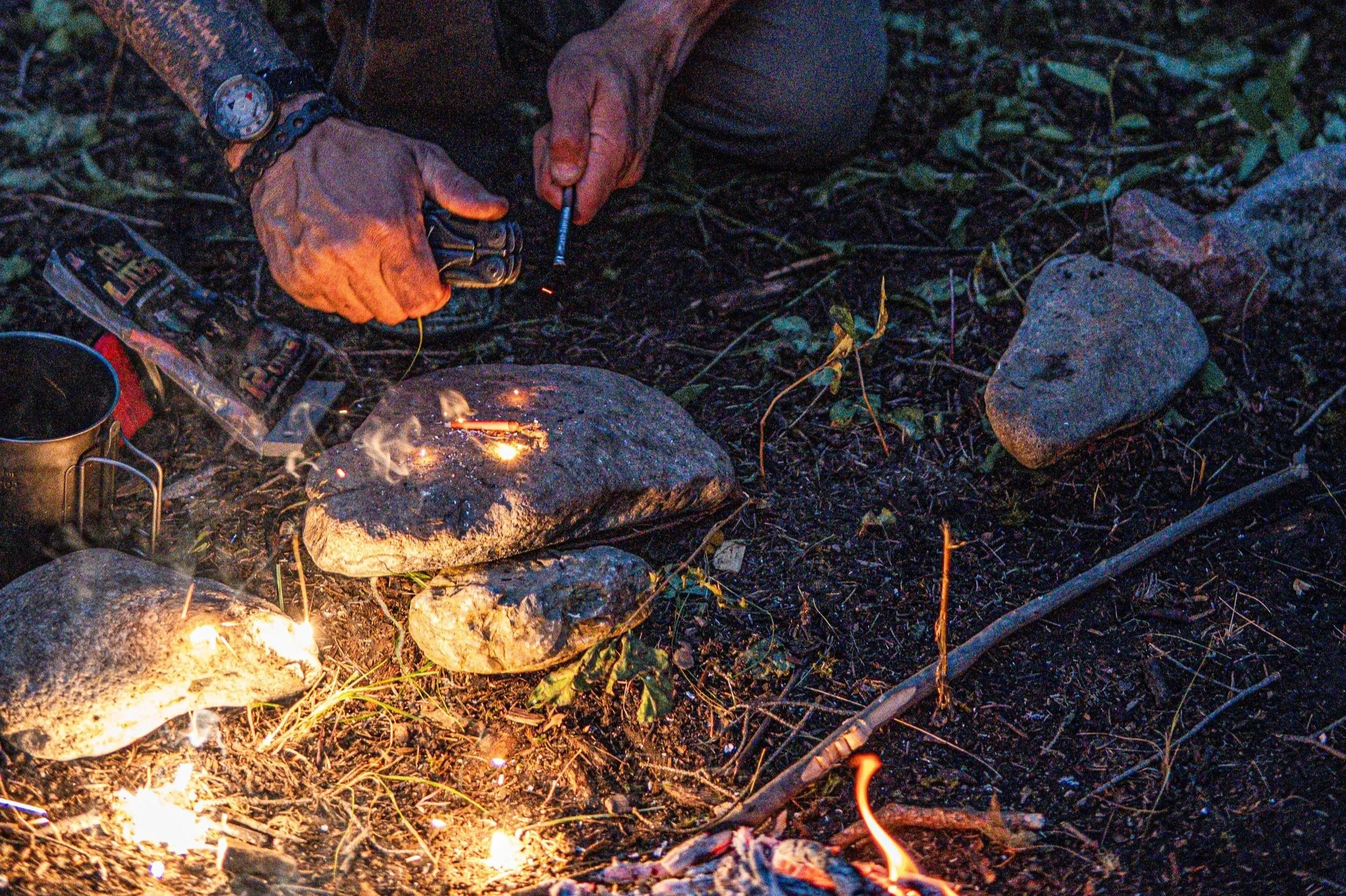 Hands holding a lighter and a metal object over small campfire on rocky ground at dusk.