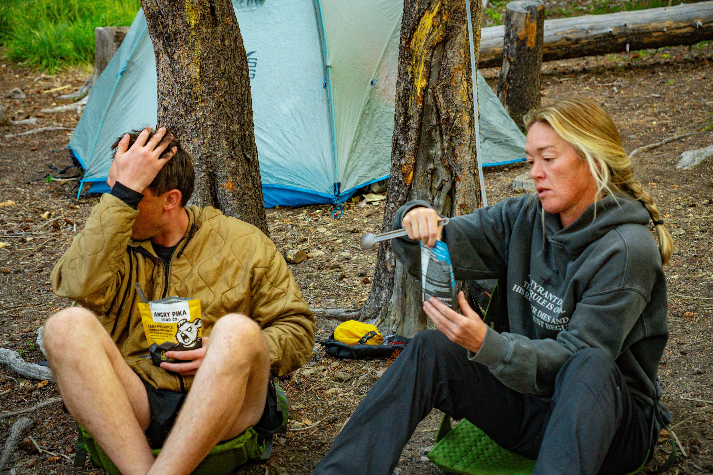 Two people sit on the ground in a forest near a tent, snack on snacks, and interact with each other.