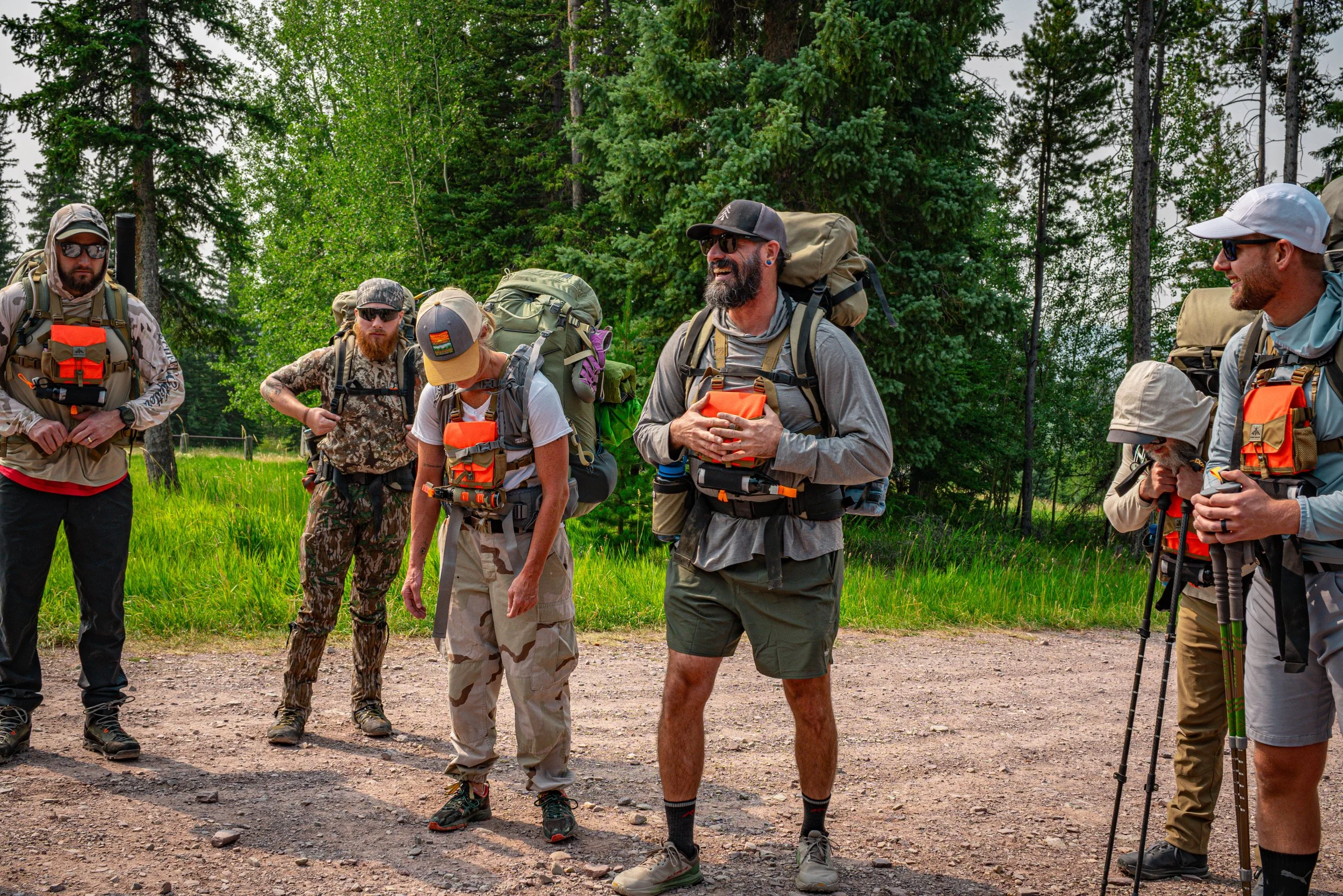 Group of hikers with backpacks walking outdoors in a forested area, some wearing sunglasses, caps, and camouflage clothing, engaging in conversation and smiling.