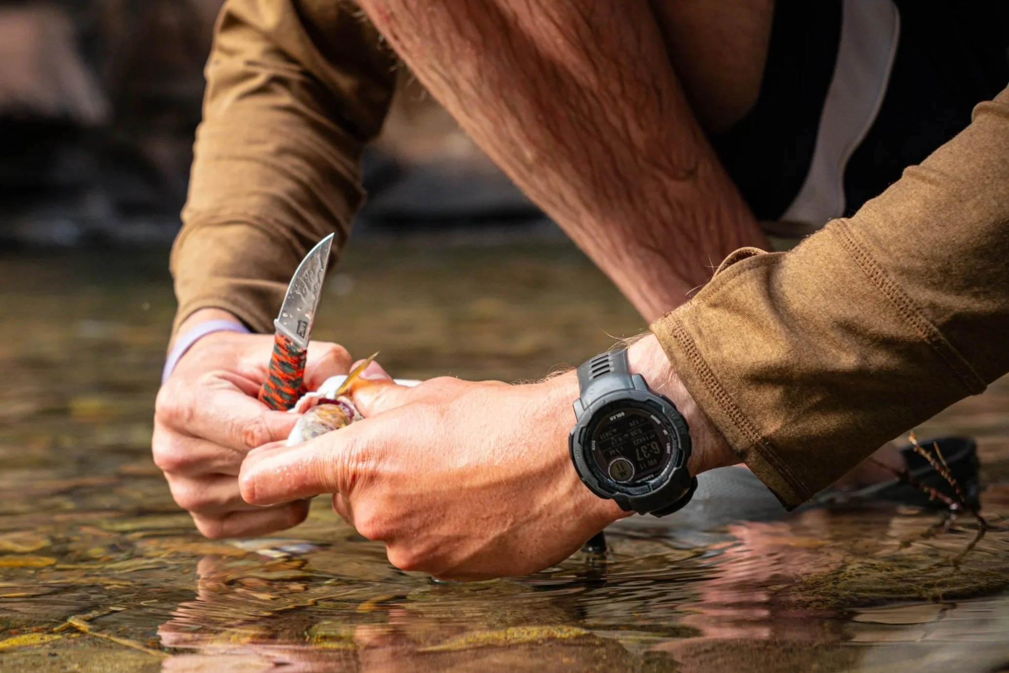 A person wearing a brown long-sleeve shirt and a black wristwatch is holding a fishing hook and handling a fish near a shallow body of water with pebbles. The person's face is not visible, and the focus is on their hands and the fish.