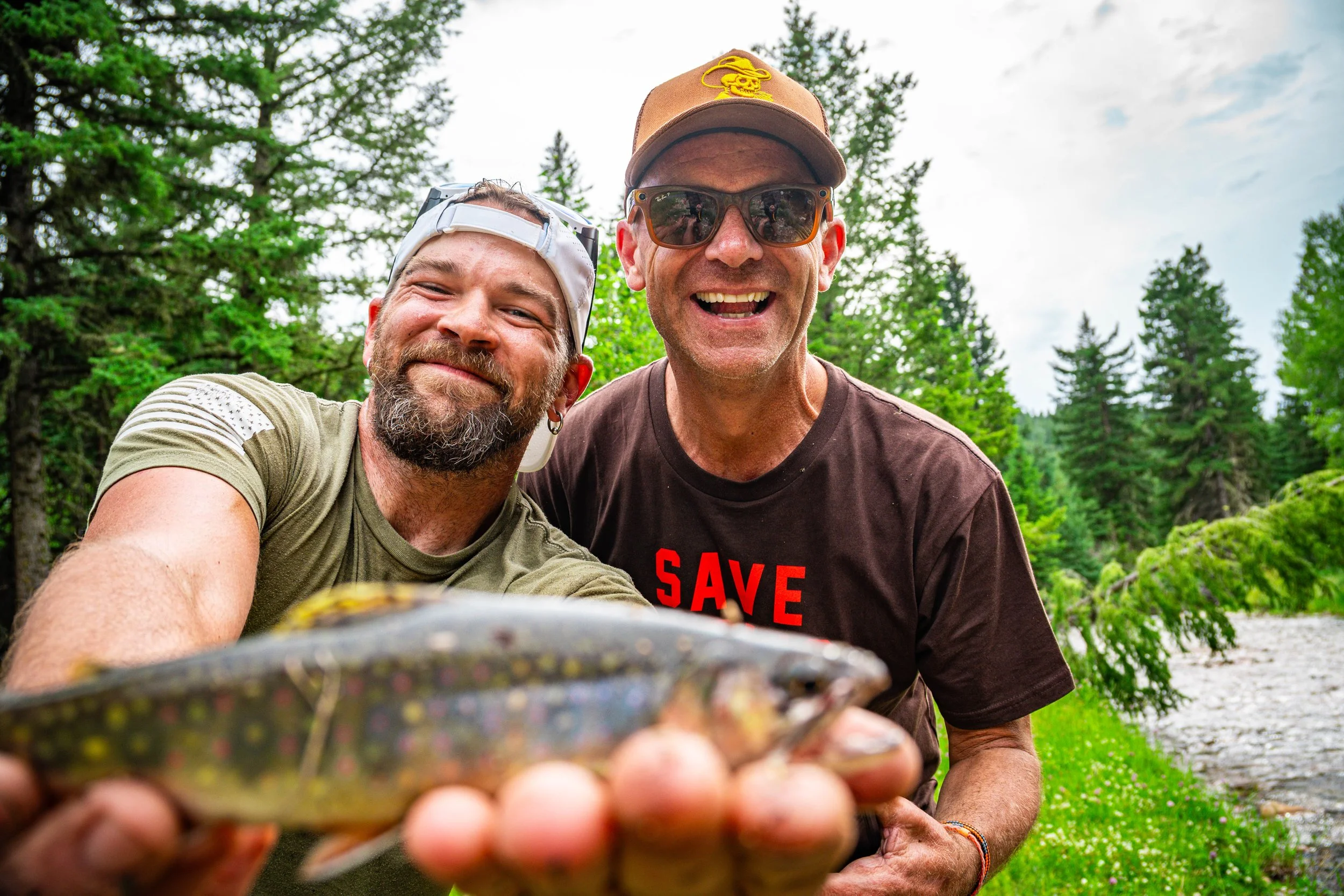 Two smiling men taking a selfie outdoors in a forested area with trees and a cloudy sky in the background.