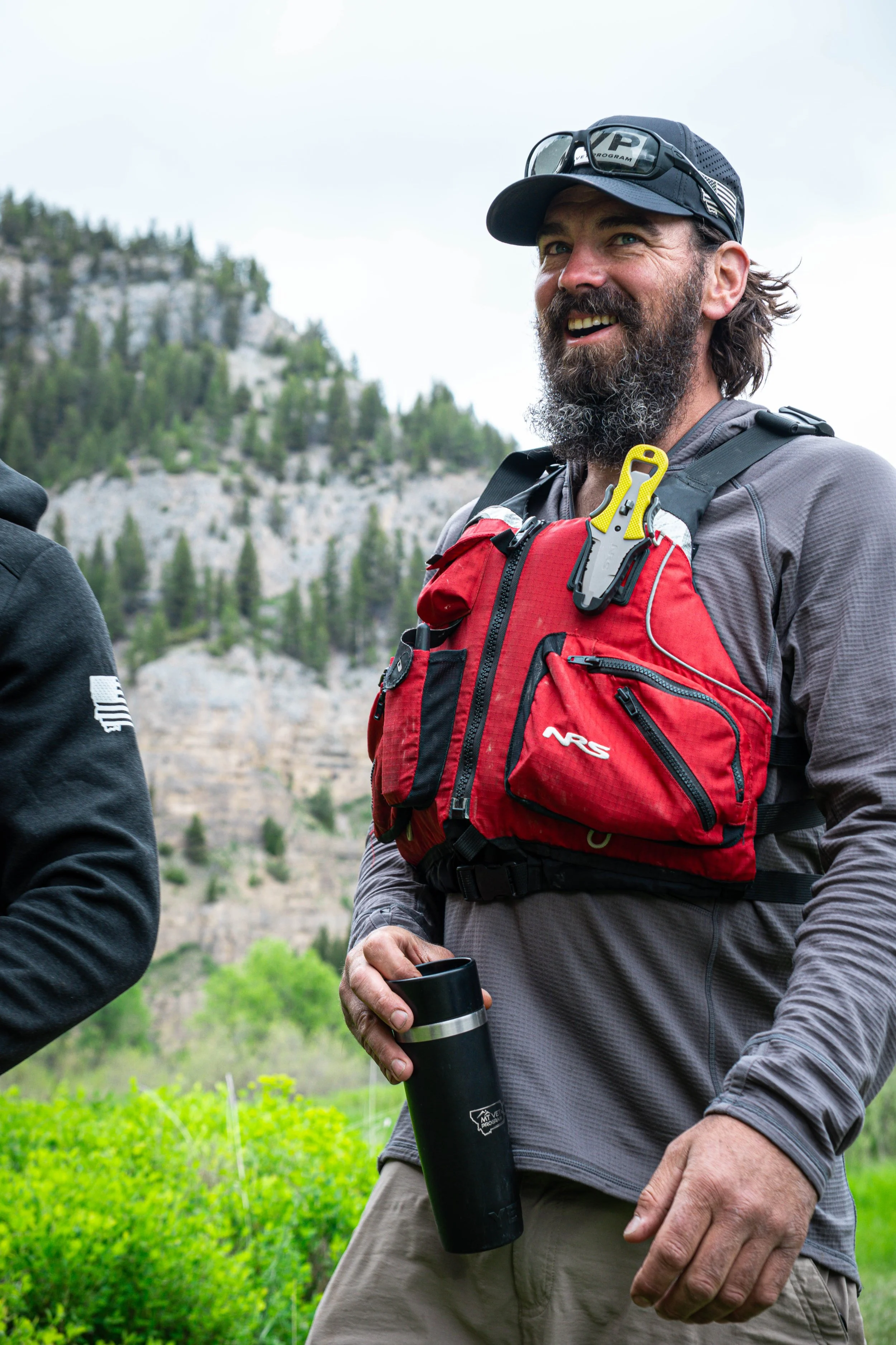 Man with beard wearing gray long-sleeve shirt, red backpack, sunglasses, and cap holding black thermos, outdoors with green bushes and mountain in background.
