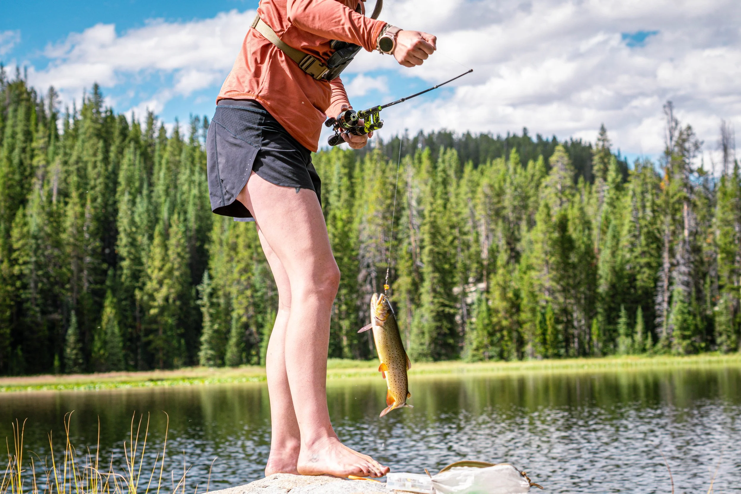 Person standing barefoot on a rock by a lake, holding a fishing rod with a fish caught on the line, with a forest and cloudy sky in the background.