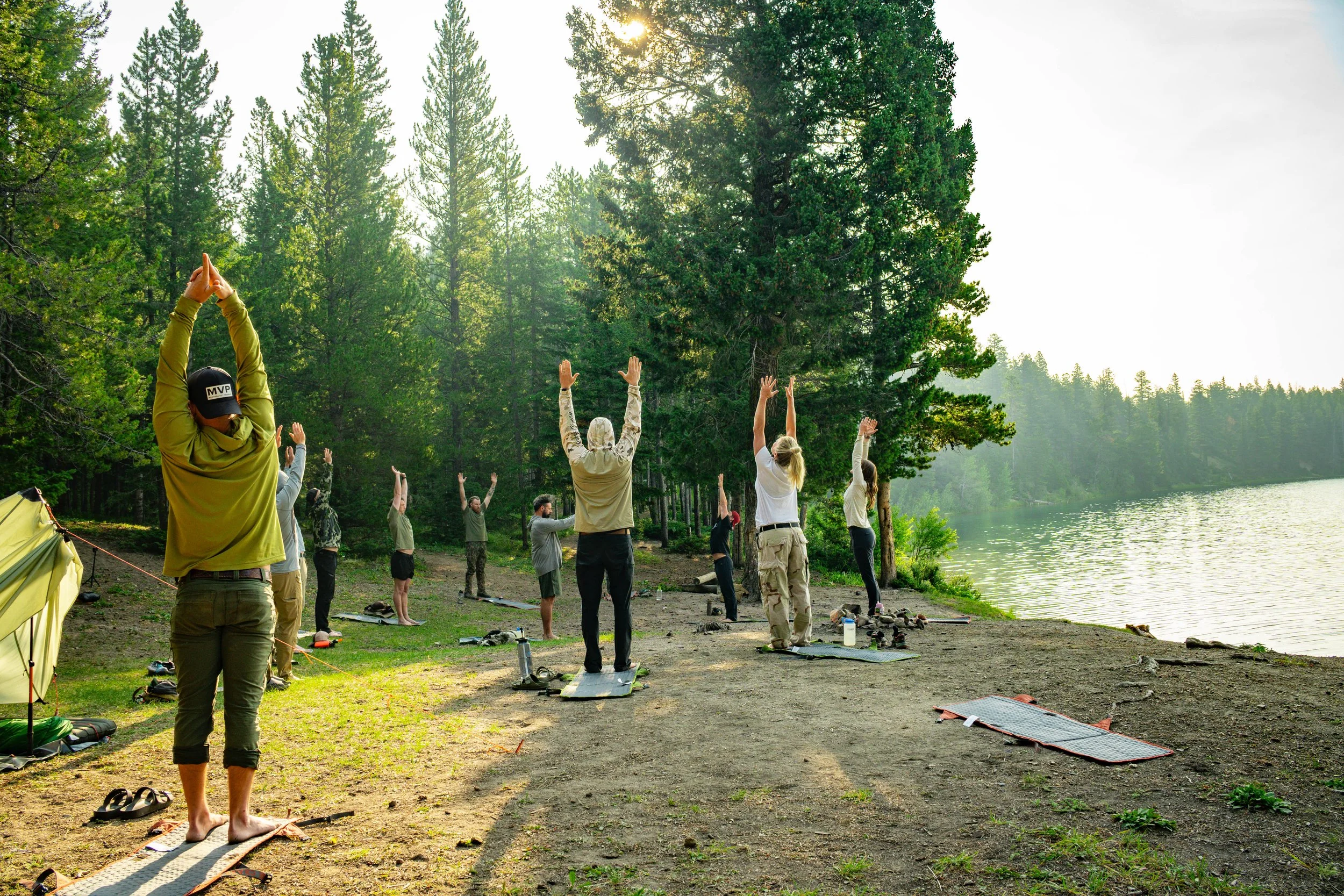 Group of people practicing yoga outdoors near a lake in a forested area during sunrise or sunset, standing on yoga mats with their hands raised above their heads.