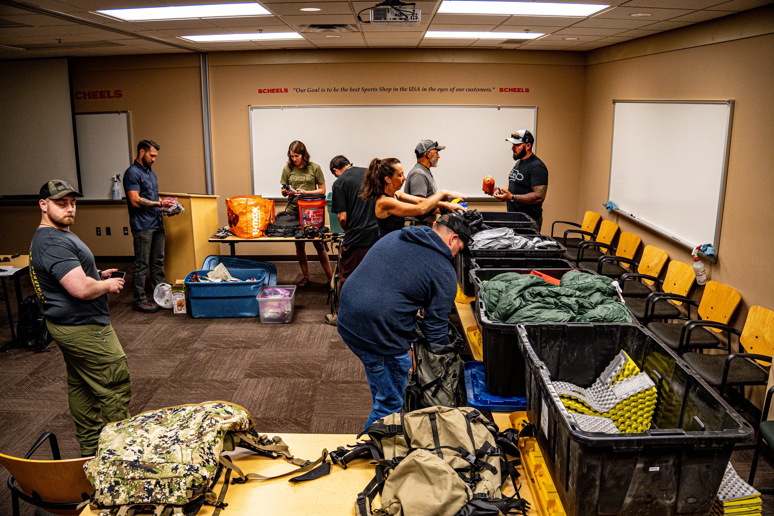 People inside a room packing gear and supplies into containers and bags, with tables and whiteboards in the background.