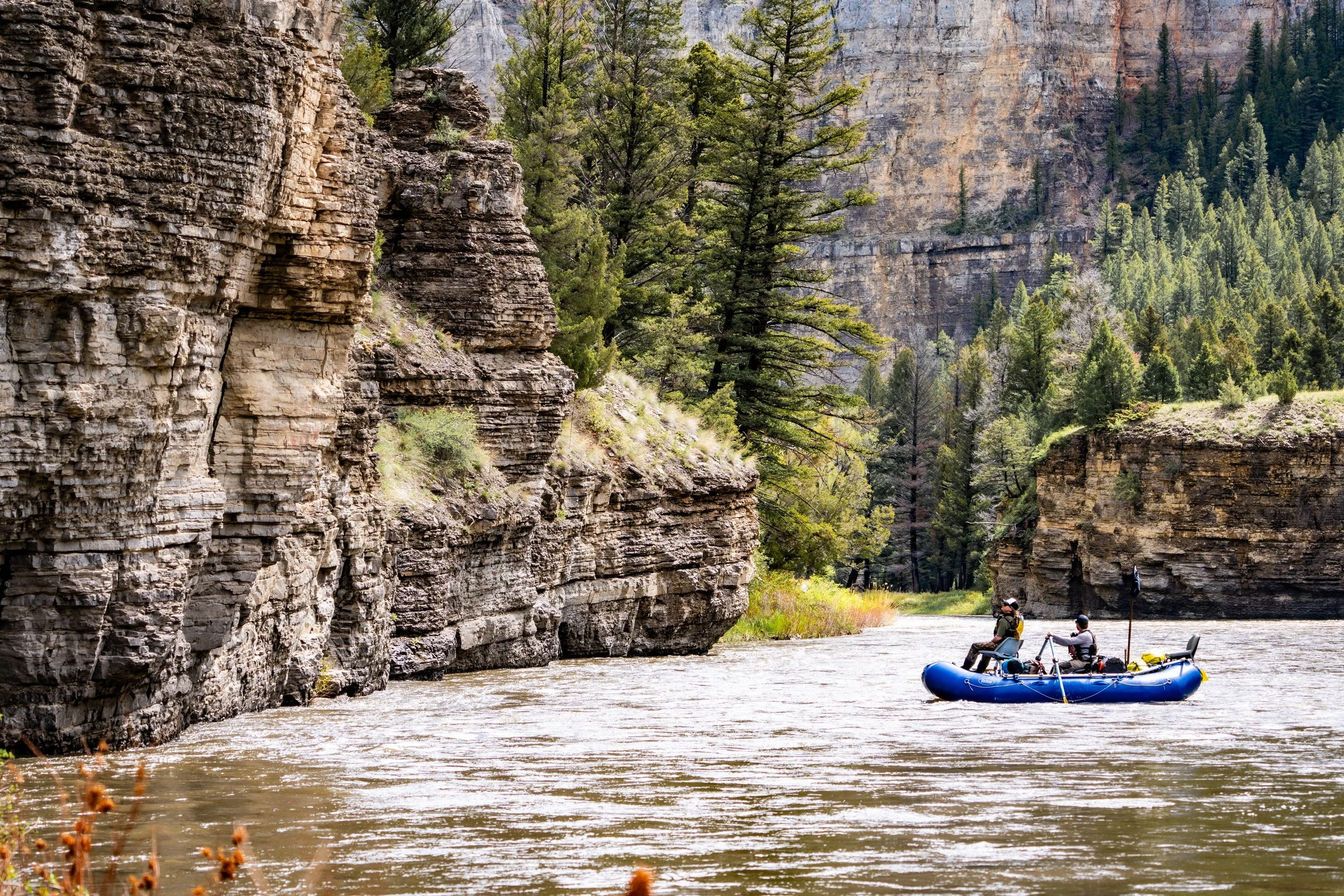People in a blue raft floating on a river with tall canyon walls and green trees in the background.