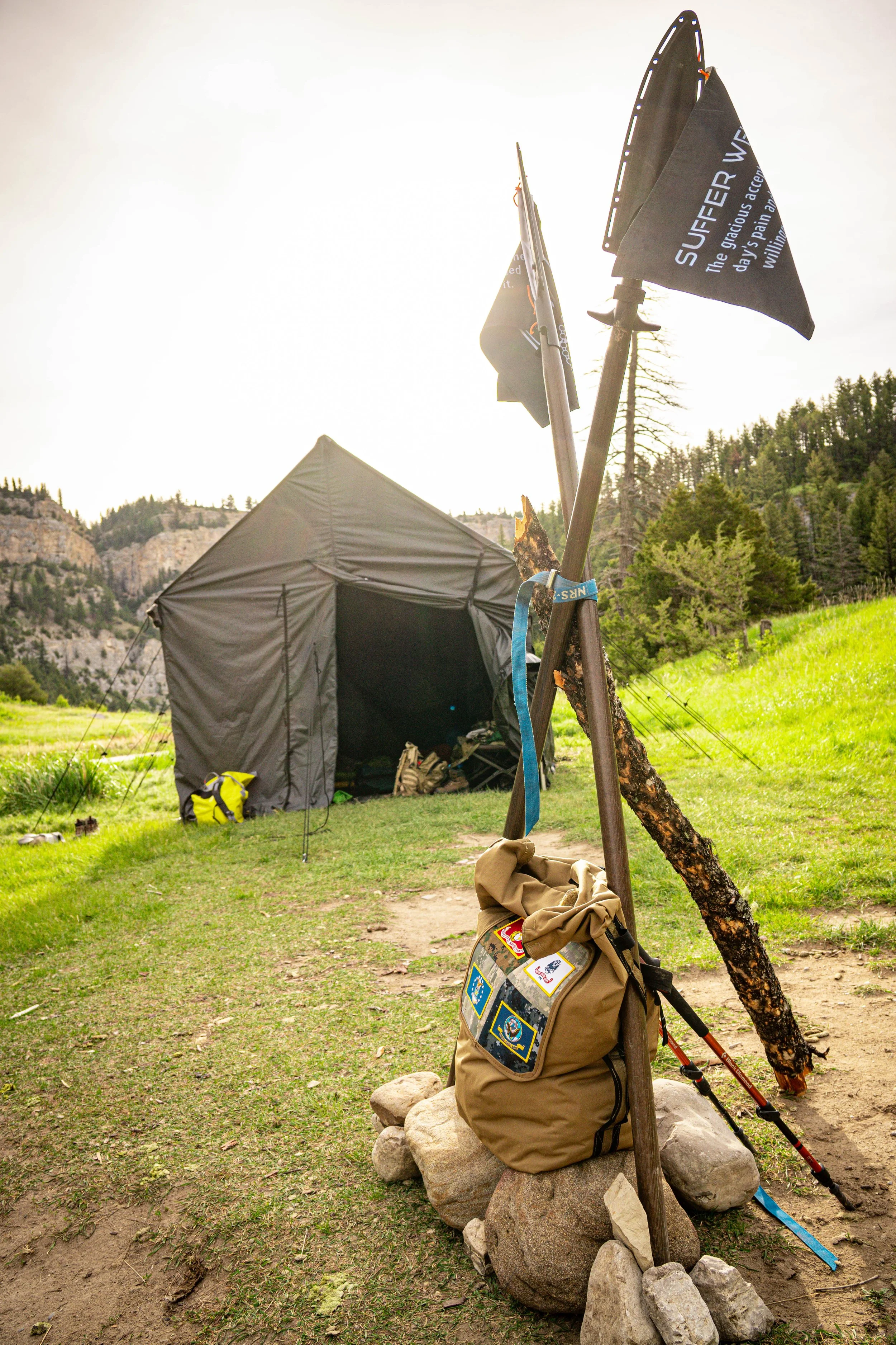 A camping scene in a grassy outdoor area with trees and hills in the background. There are two black flags with white text on the poles, a gray tent, and a PIG EGG sitting on rocks and leaning again