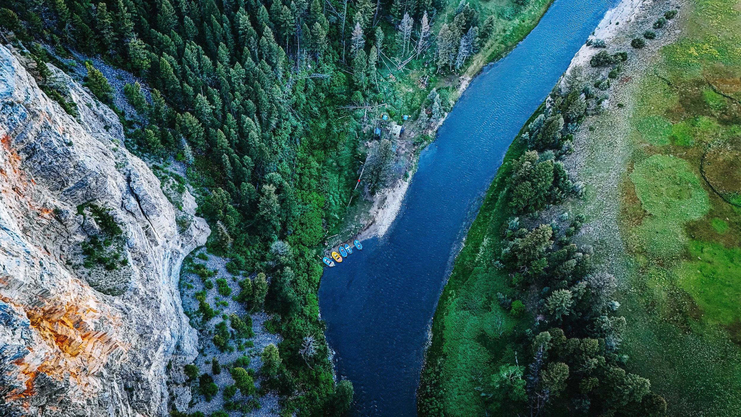 Aerial view of a river flowing through a forested valley with boats docked along the shoreline and rocky cliffs on one side.