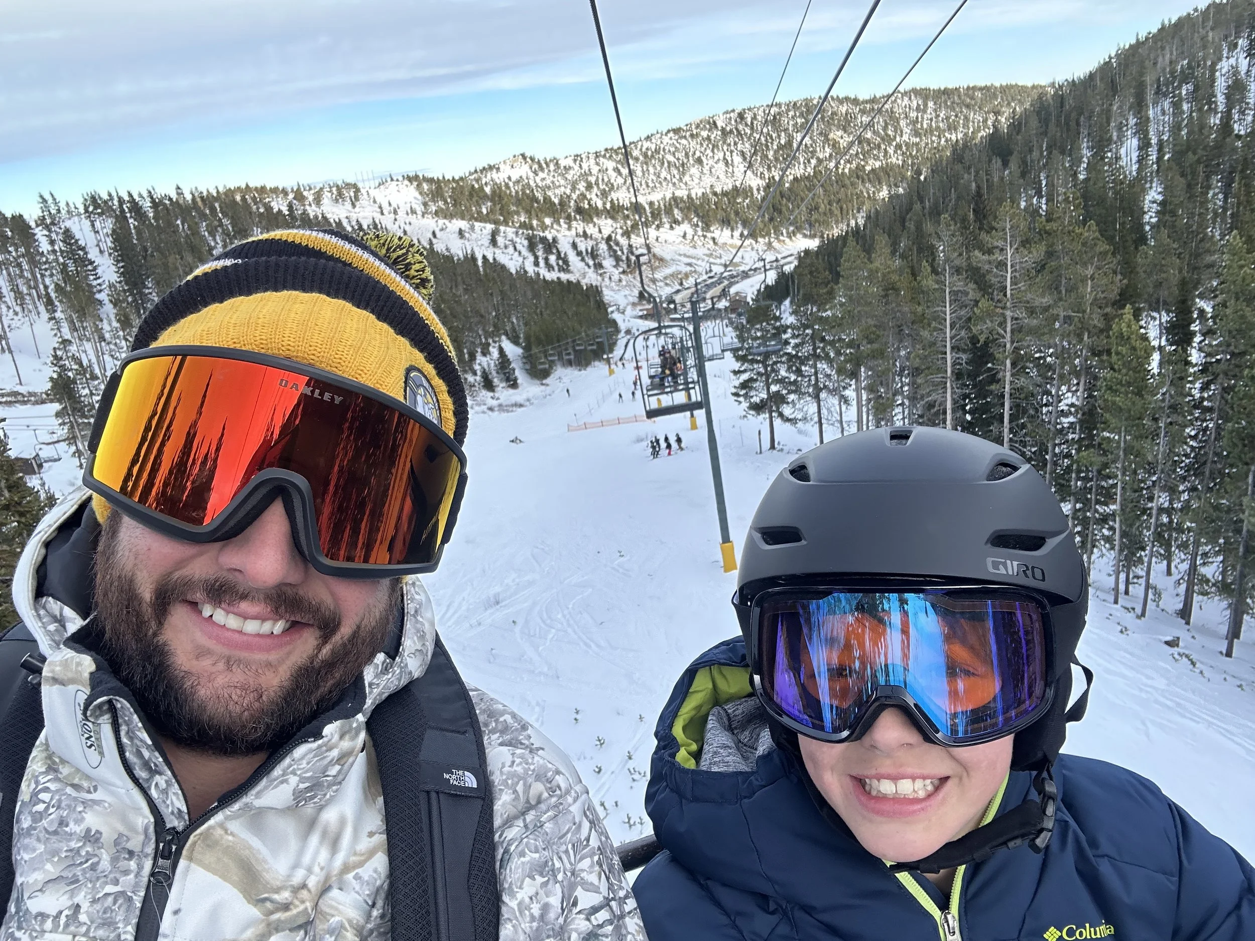 Two people in winter ski gear taking a selfie on a snowy mountain with ski lifts, trees, and other skiers in the background.