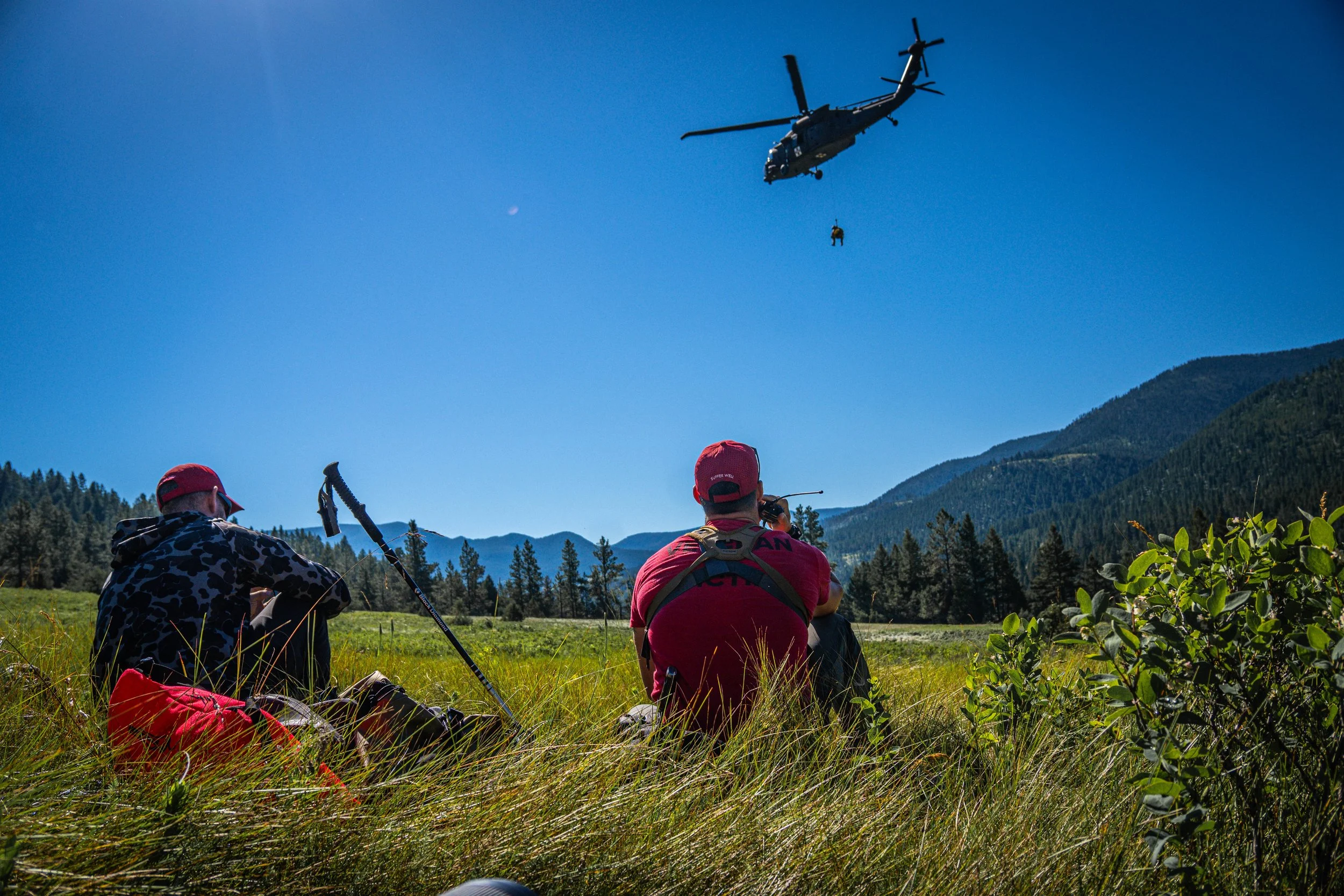 Three people sitting on grass outdoors, watching a helicopter fly overhead with a person hanging from its rope in a mountainous area.