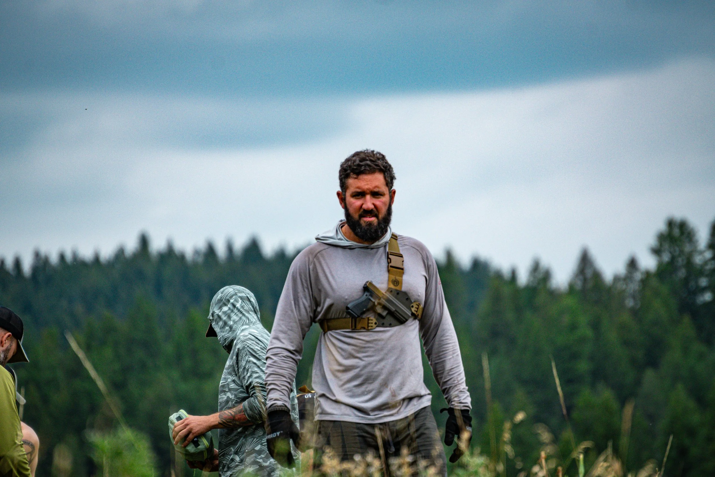 A man with a beard and curly hair, wearing a gray long-sleeve shirt and tactical gear, walking outdoors in a natural setting with trees in the background.
