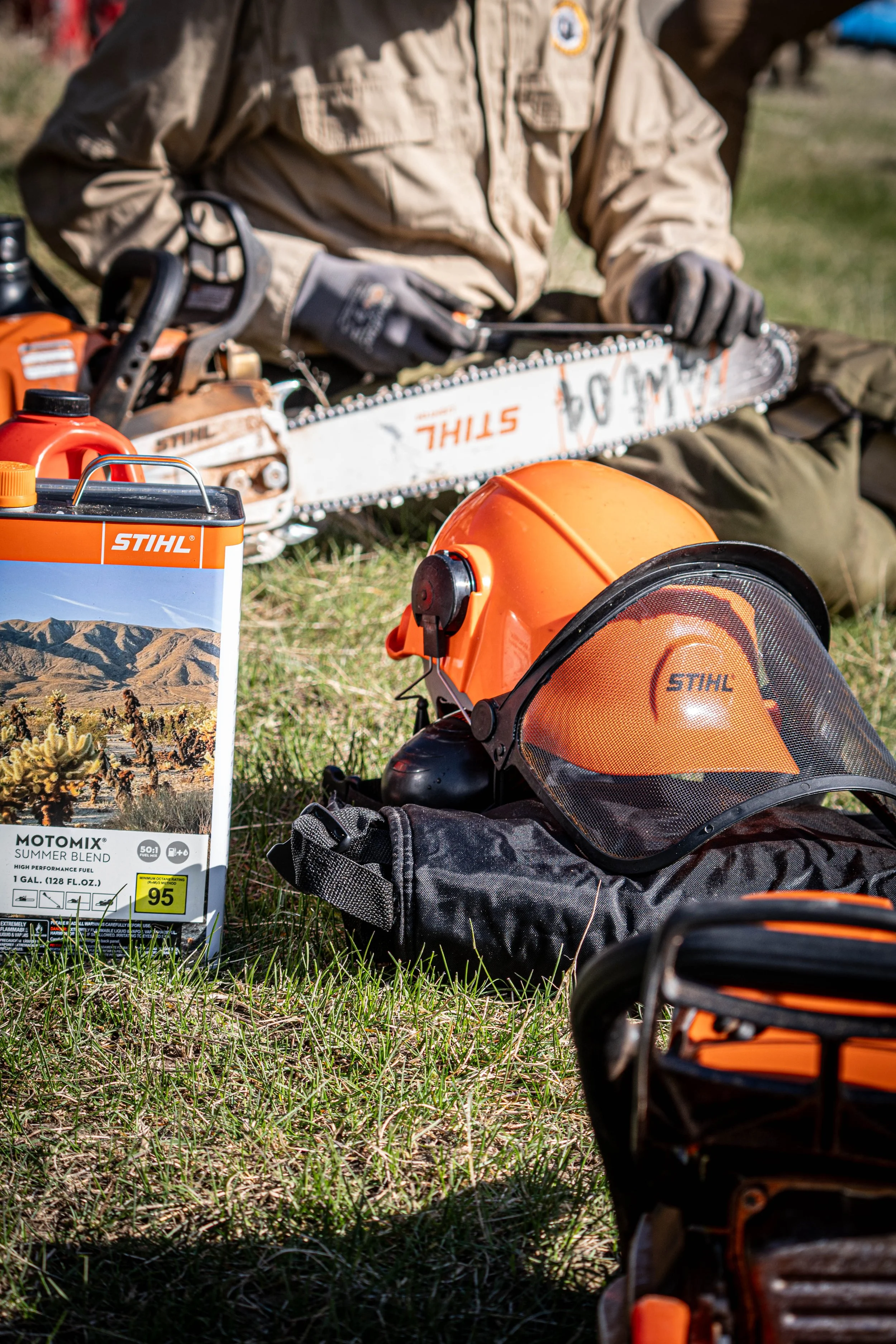 Person wearing protective gear holds a chainsaw outdoors, with safety helmet nearby, and fuel and chainsaw oil containers on grass.