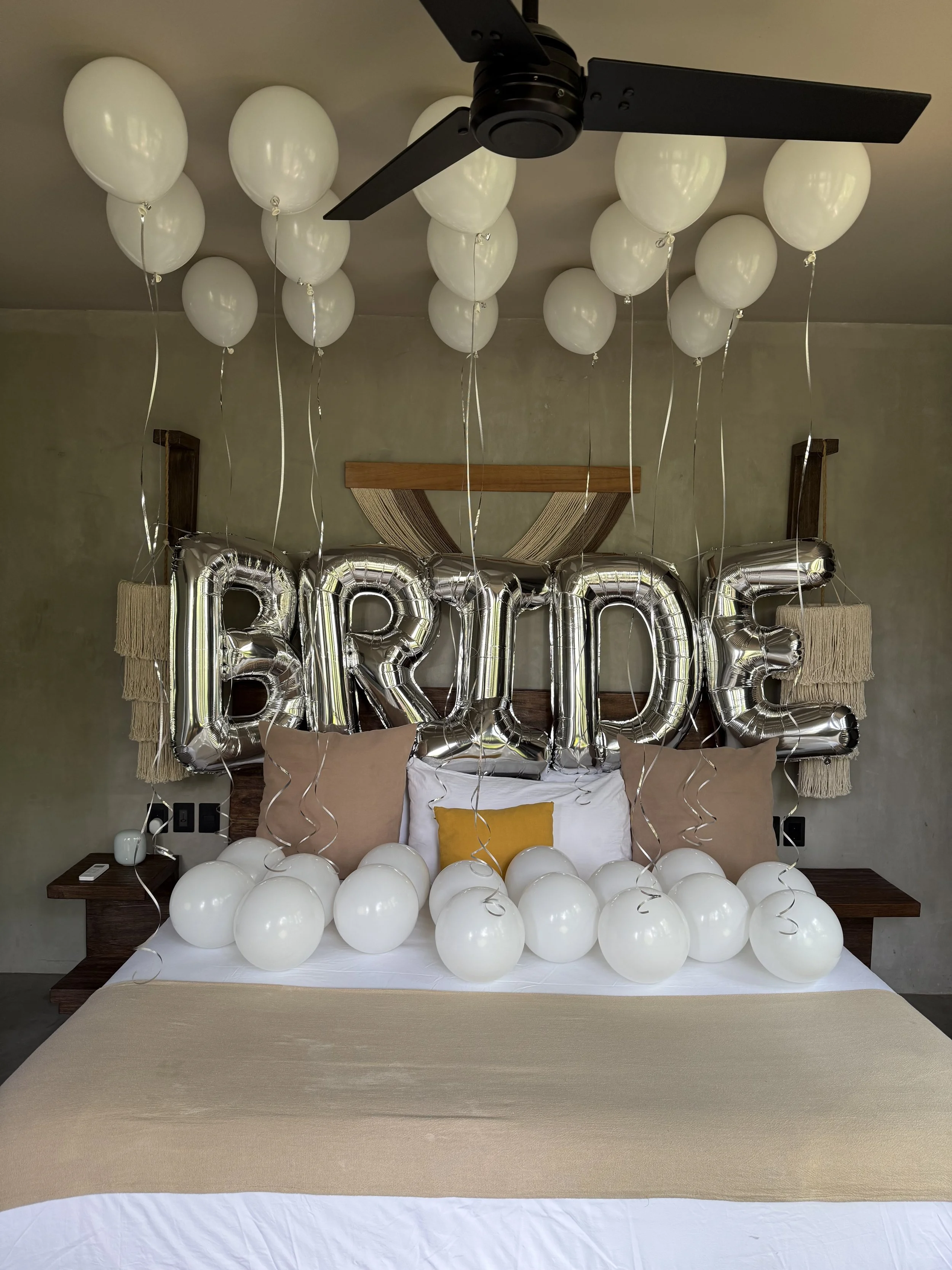 Decorated bed with white balloons on the headboard and around the bed, silver "HAPPY" balloons hanging above, and a ceiling fan.
