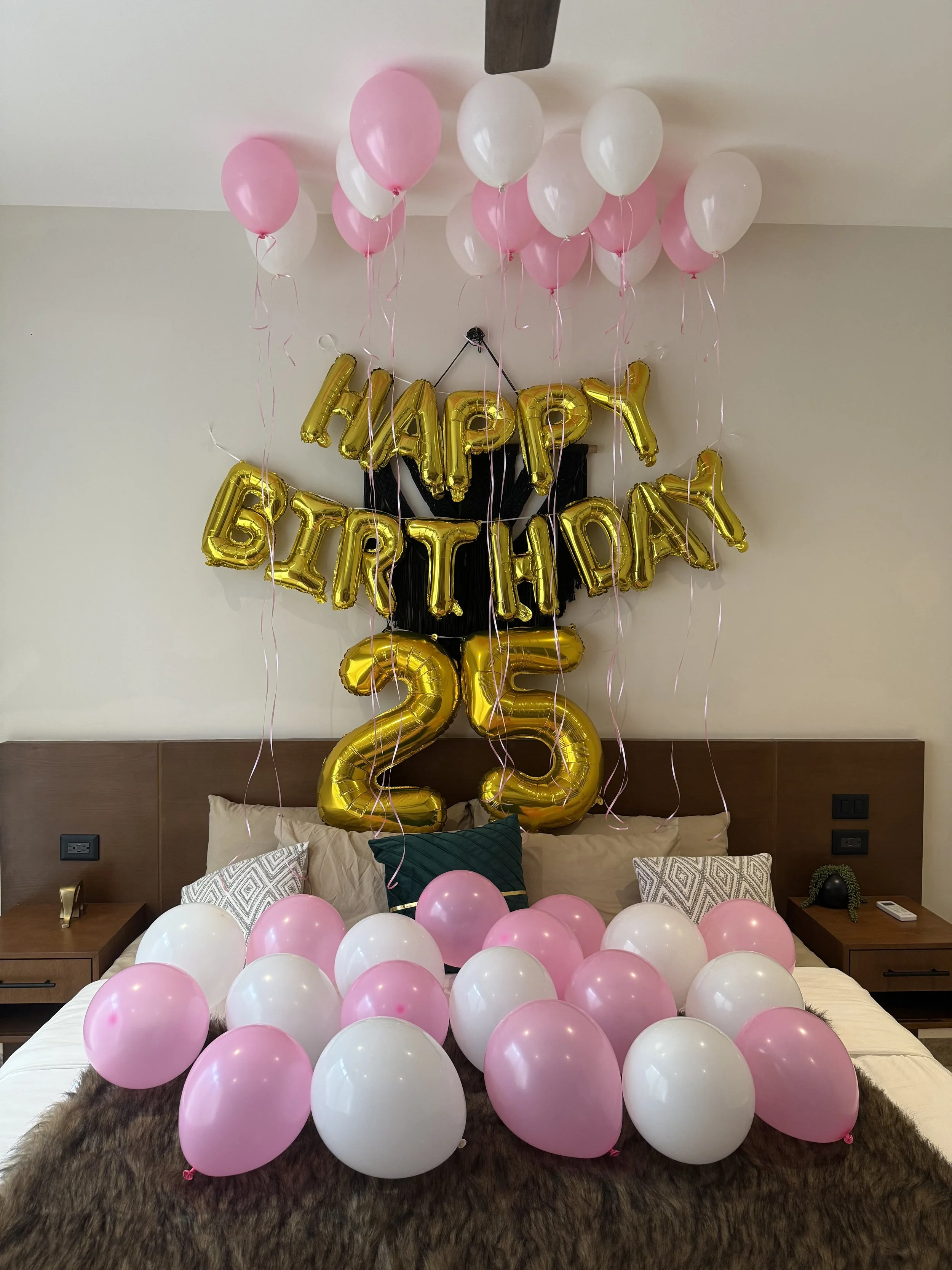 Decorated bedroom with 'Happy Birthday' balloon banner, large gold balloons forming the number 25, and pink and white balloons on the bed and ceiling for a birthday celebration.