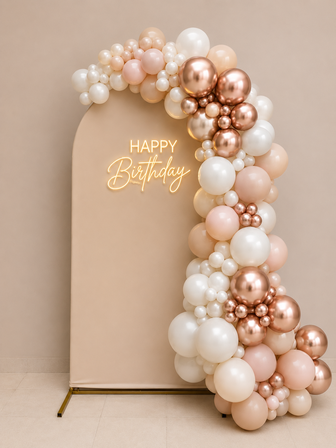 A beige arch-shaped backdrop decorated with pink, white, and metallic rose gold balloons, with a neon sign reading 'HAPPY Birthday' on the left side.