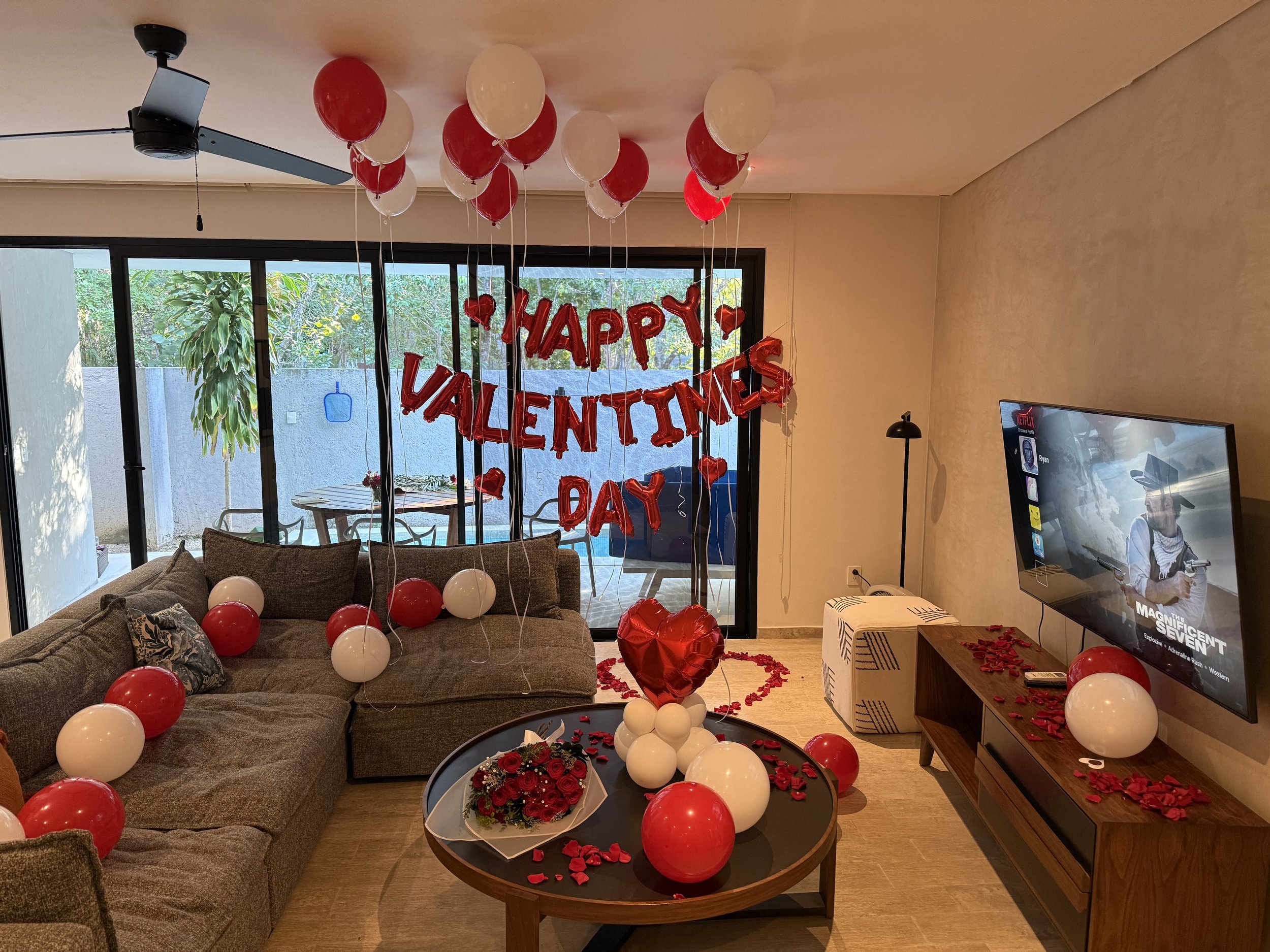 Living room decorated for Valentine's Day with red and white balloons, a 'Happy Valentine's Day' banner, heart-shaped balloon, and red rose petals on the table and floor. There's a sofa with pillows, a TV, and a balcony visible through glass sliding 