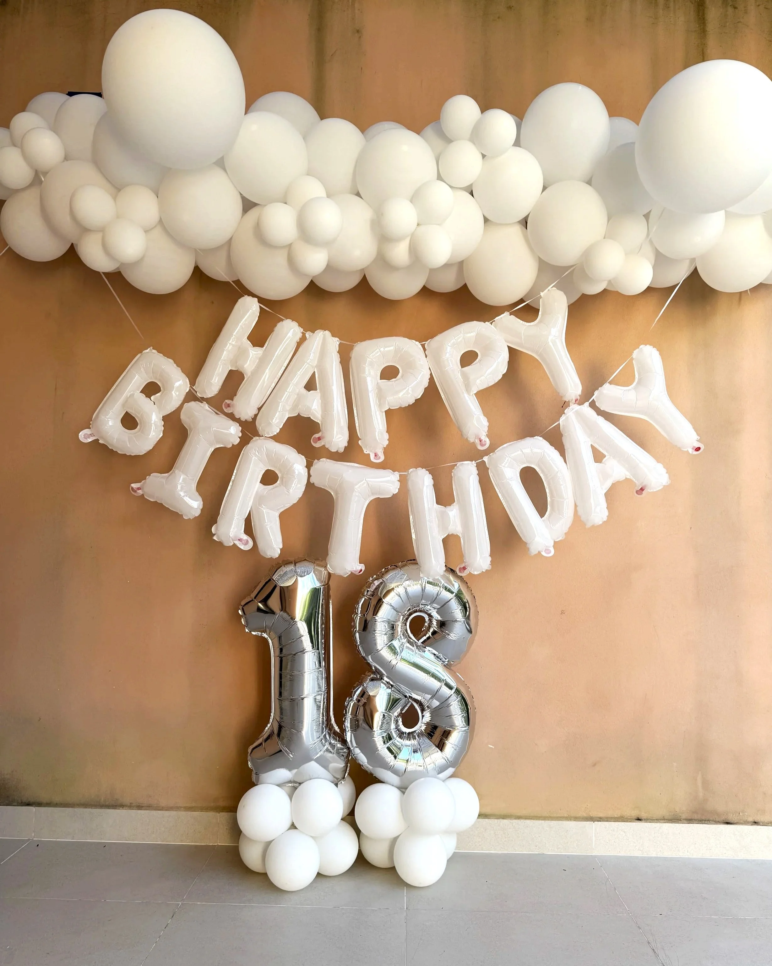 Celebration wall decoration with white balloons, metallic number balloons '18', and white letter balloons spelling 'HAPPY BIRTHDAY' against a brown wall.
