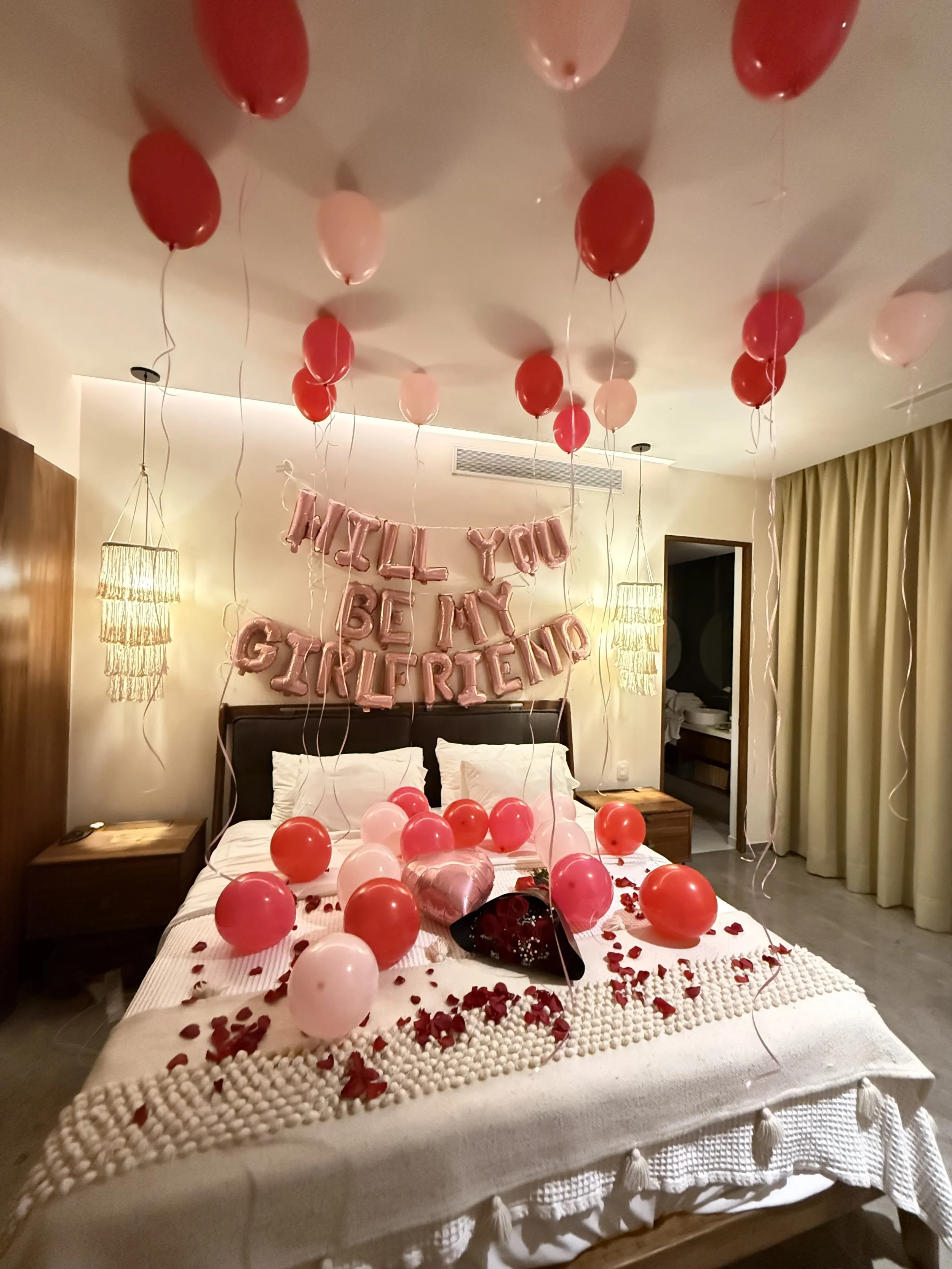 A decorated bedroom with pink and red balloons, rose petals, a heart-shaped balloon, and a balloon banner that reads "Will You Be My Girlfriend." The bed has white bedding and is surrounded by romantic decorations for a proposal.