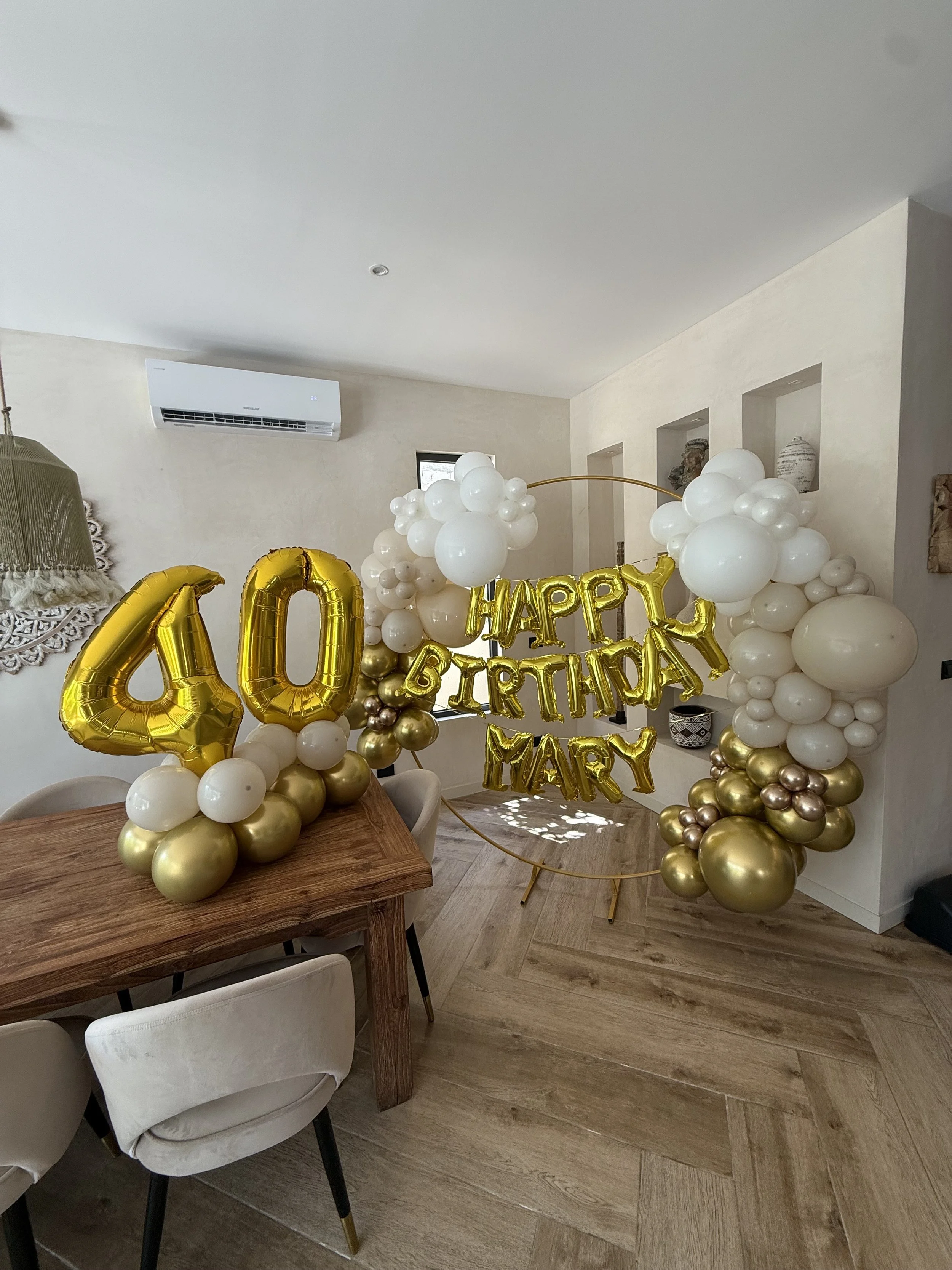 Decorations for a 40th birthday celebration, featuring large gold balloons displaying the number '40', and additional gold and white balloons with a gold 'Happy Birthday Mary' sign, set up in a dining room with a wooden table and beige chairs.