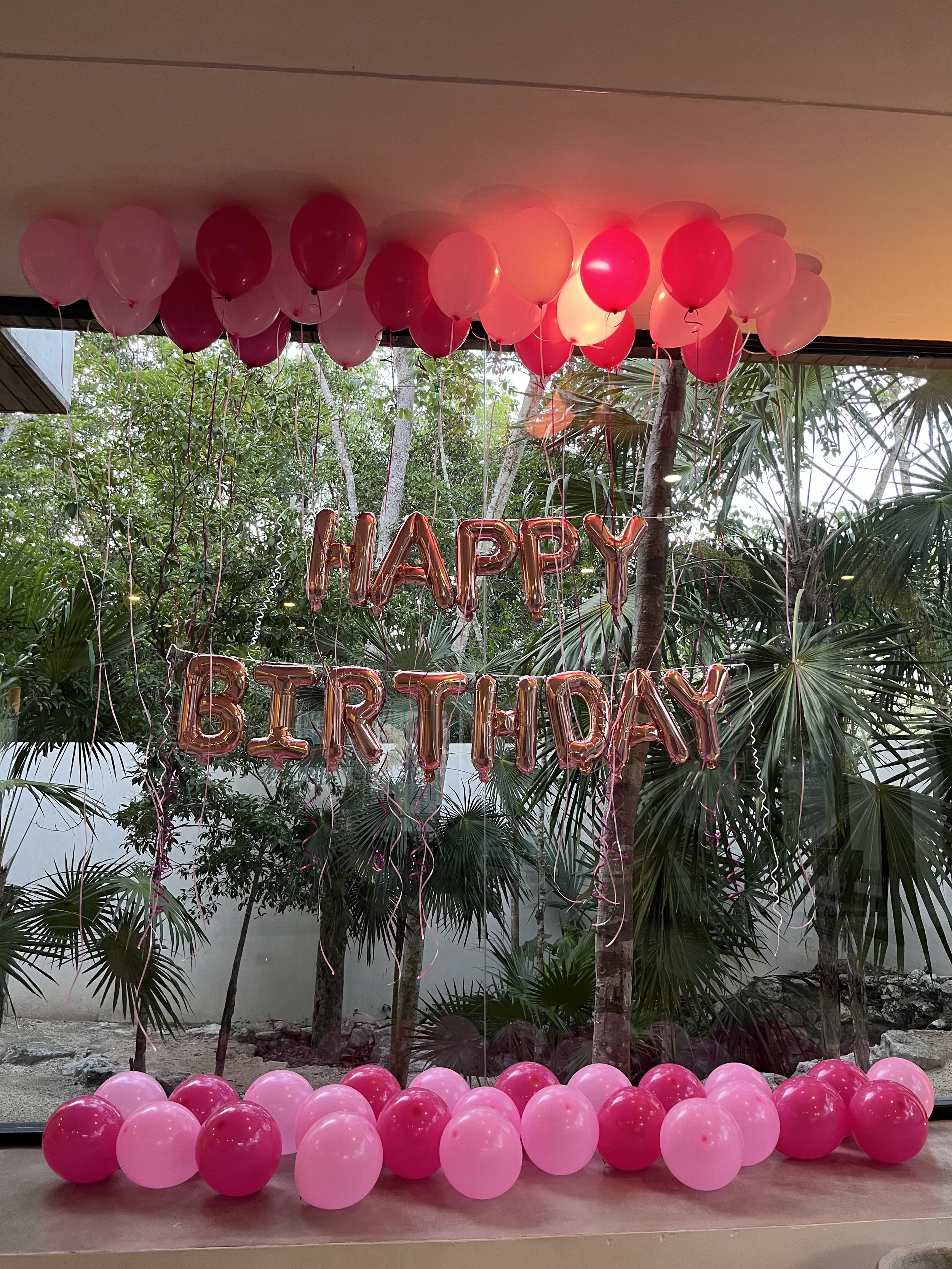 Pink and red balloons arranged at a birthday celebration with a 'Happy Birthday' balloon banner in the background, set outdoors with trees and greenery visible behind.