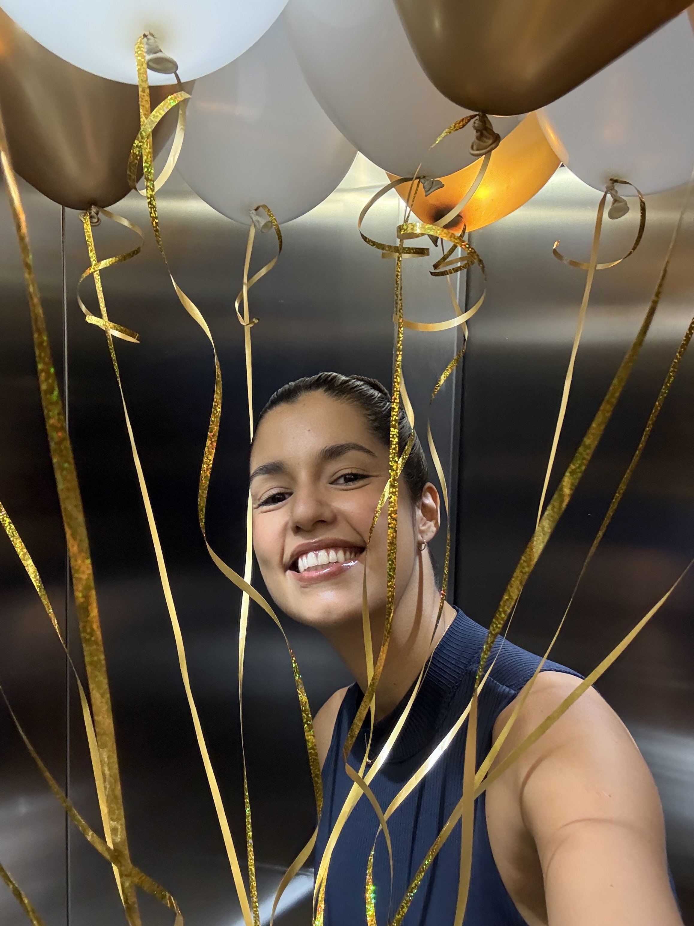 A woman taking a selfie surrounded by gold, silver, and white balloons with gold ribbons, smiling in an elevator.