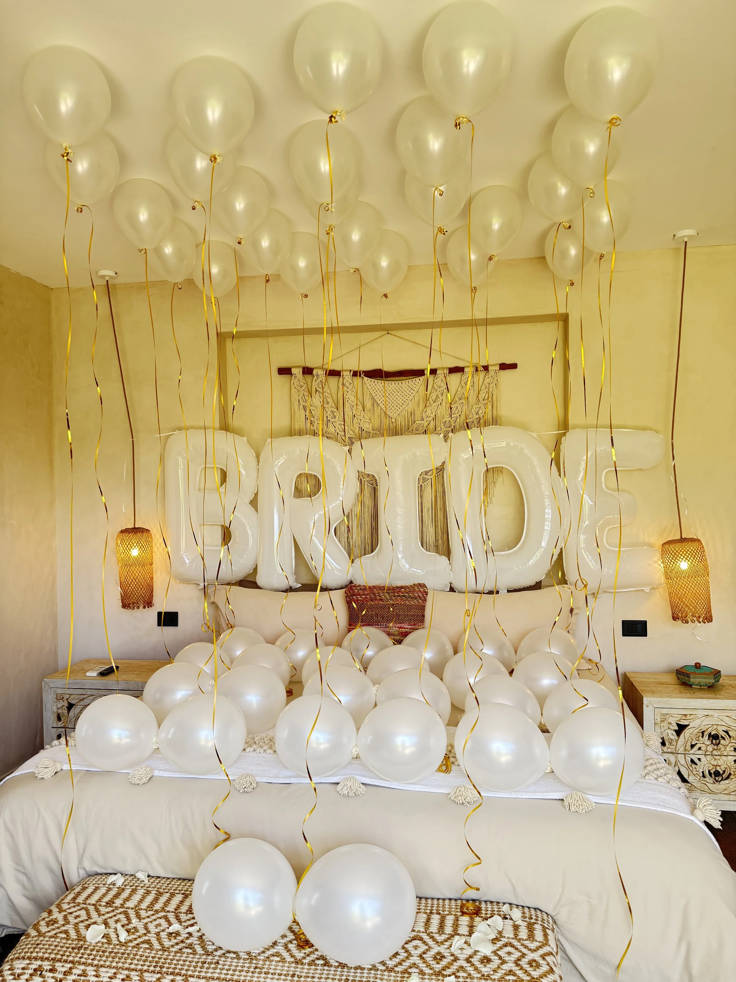 Decorated bedroom with white balloons, gold ribbons, large white balloons spelling out 'BABY', warm lighting, and cozy furnishings.