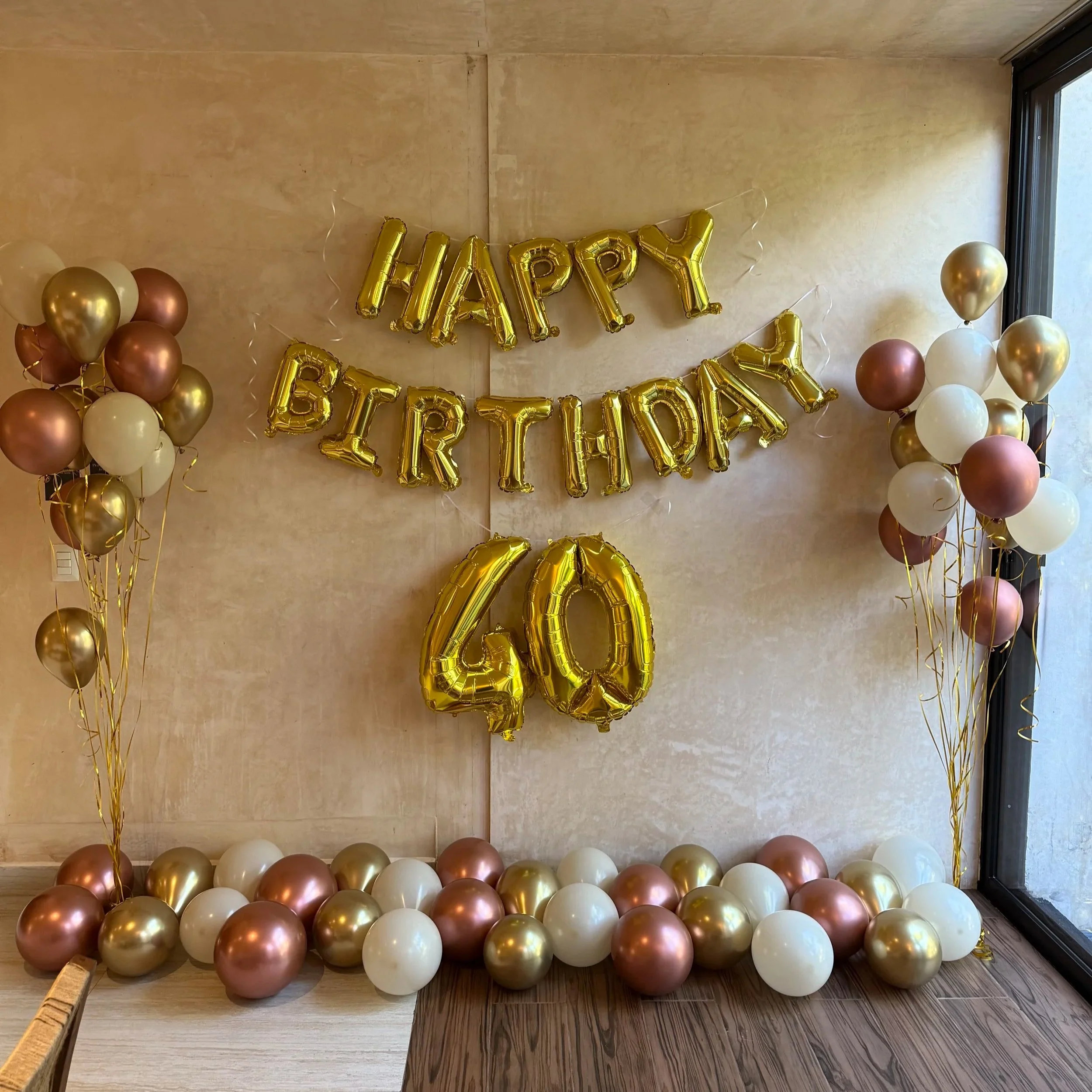 Decorative birthday setup with gold and white balloons, balloons spelling out 'HAPPY BIRTHDAY' in gold, and a large gold '40' balloon in the center. The background is a beige wall near a large window.