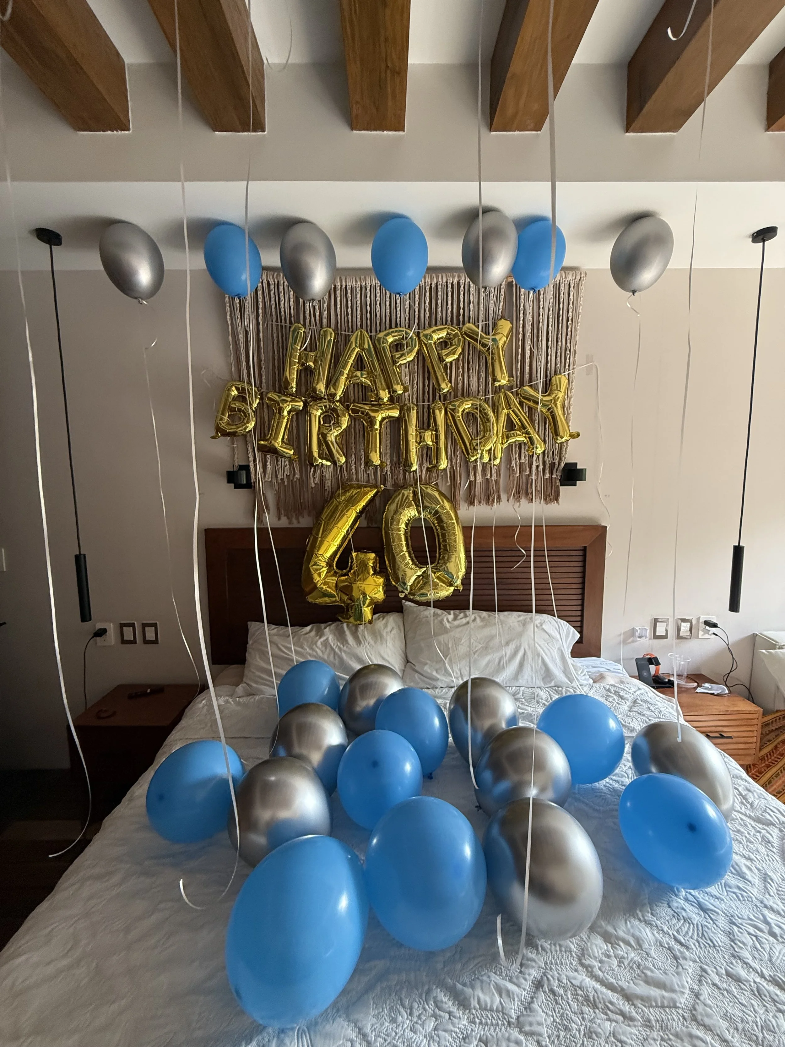 Decorated bedroom with blue, silver, and gold balloons, including large gold helium balloons spelling out 'HAPPY BIRTHDAY' and the number 40, on a bed with white bedding, celebrating a 40th birthday.