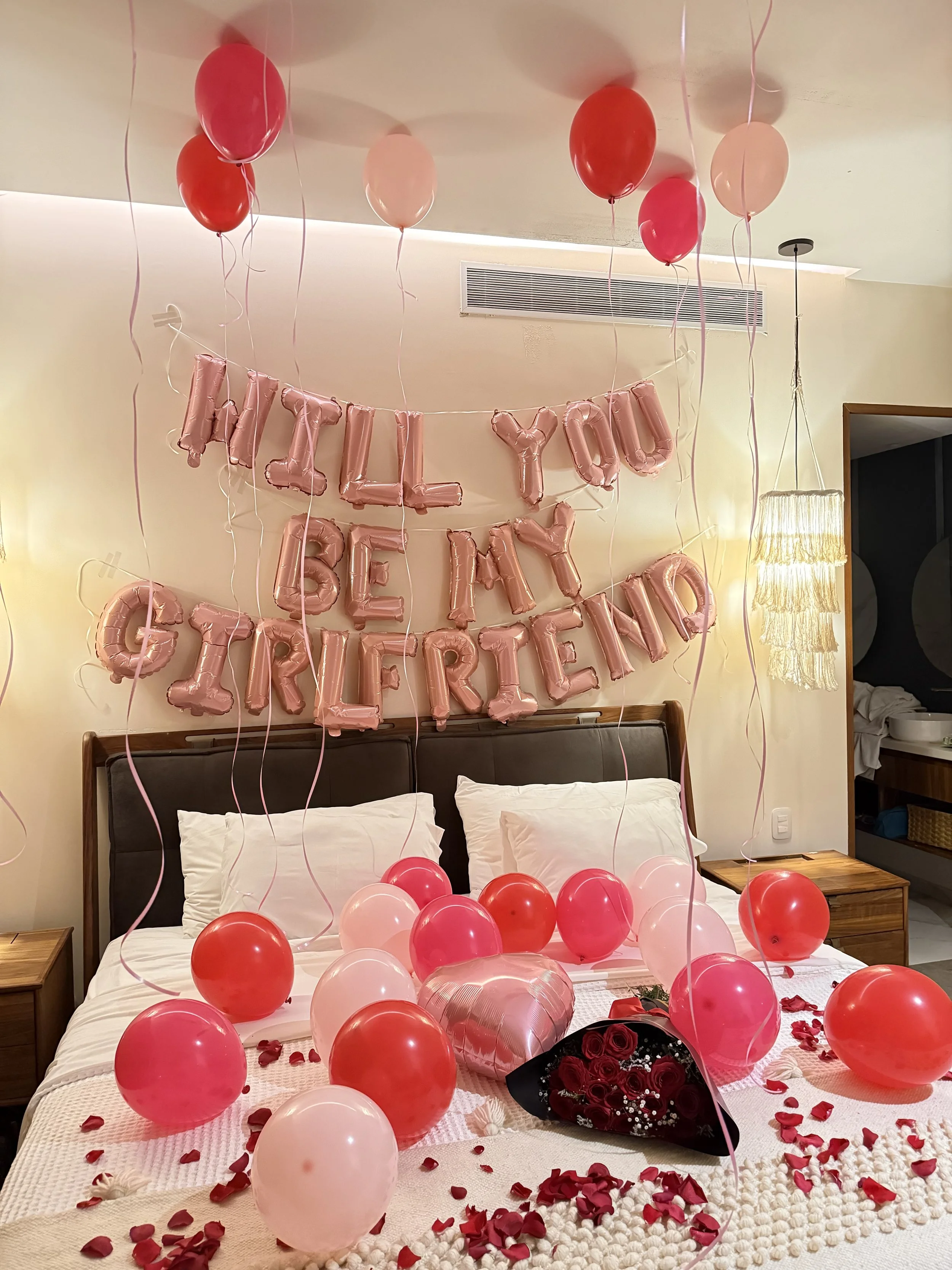 Decorated bedroom with pink and red balloons, a balloon banner saying 'Will You Be My Giraffe Rriend', flower bouquet, and rose petals on the bed for a birthday celebration.