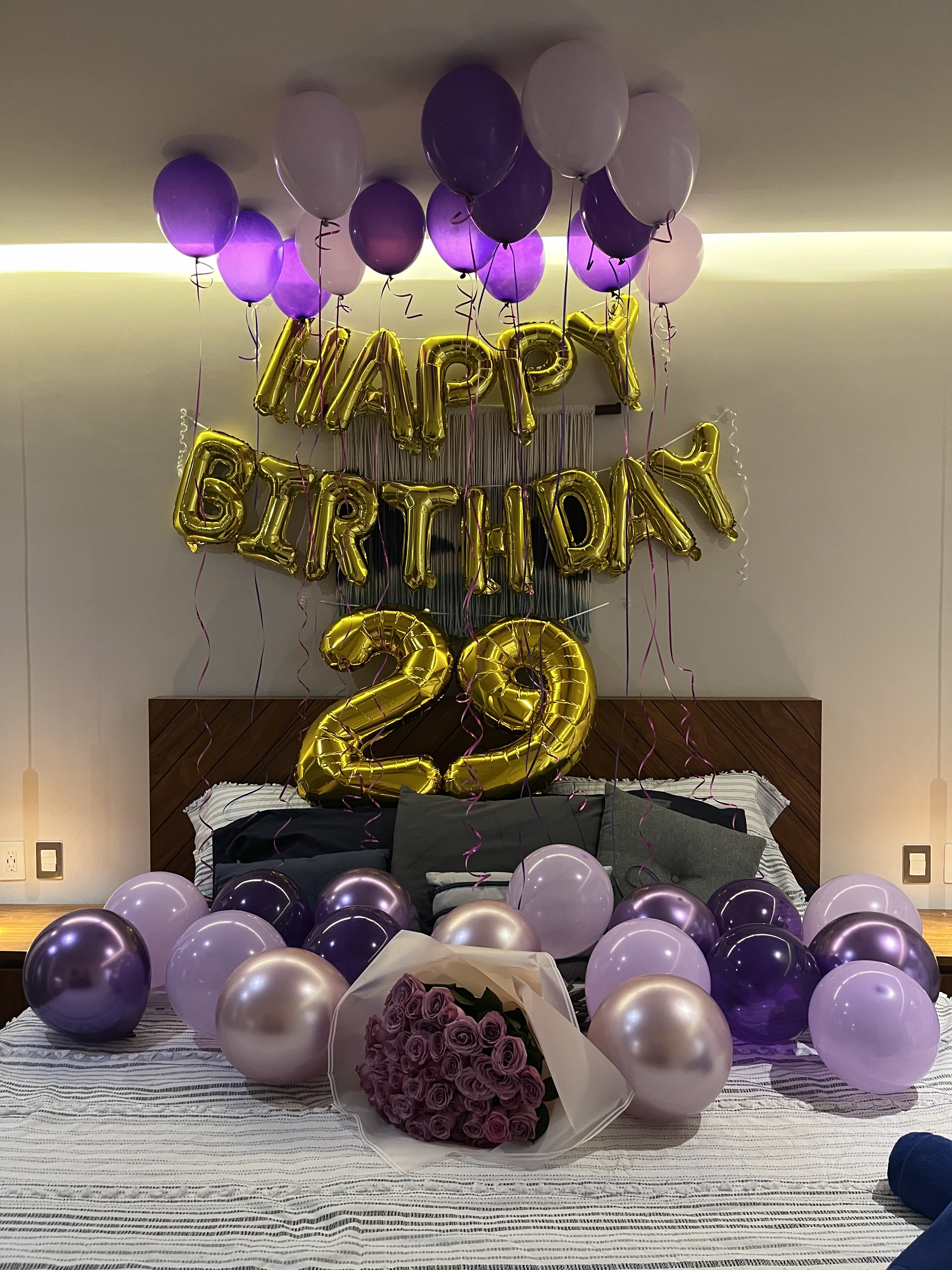 Decorated bedroom with pink and purple balloons, gold 'Happy Birthday' and the number '25' balloons, a bouquet of purple roses, and a striped bedspread.