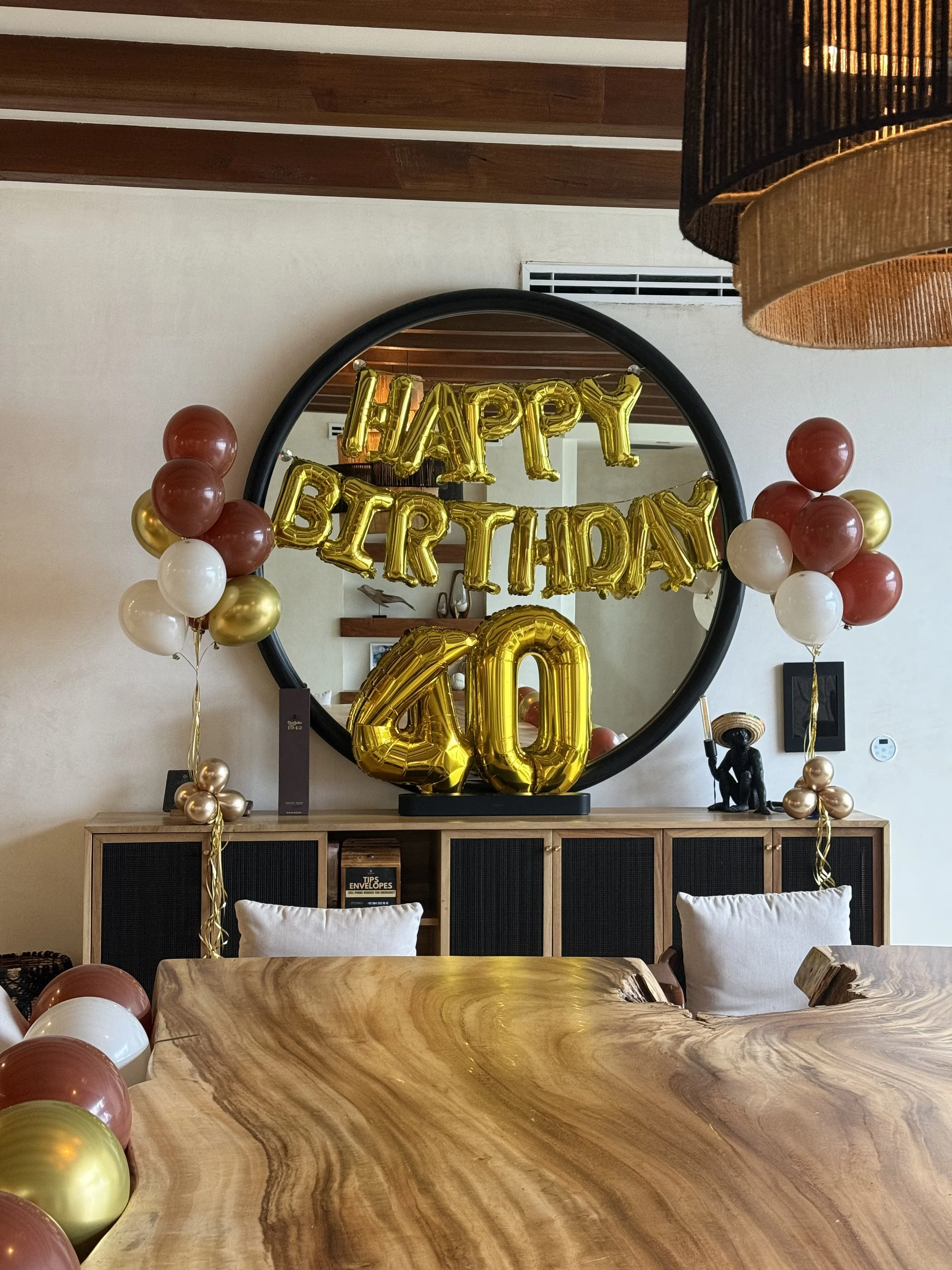 Decorations for a 40th birthday celebration, including gold balloons spelling out "Happy Birthday" and the number "40," surrounded by balloons in gold, white, and maroon, with a large mirror reflecting the decorations in a living room.