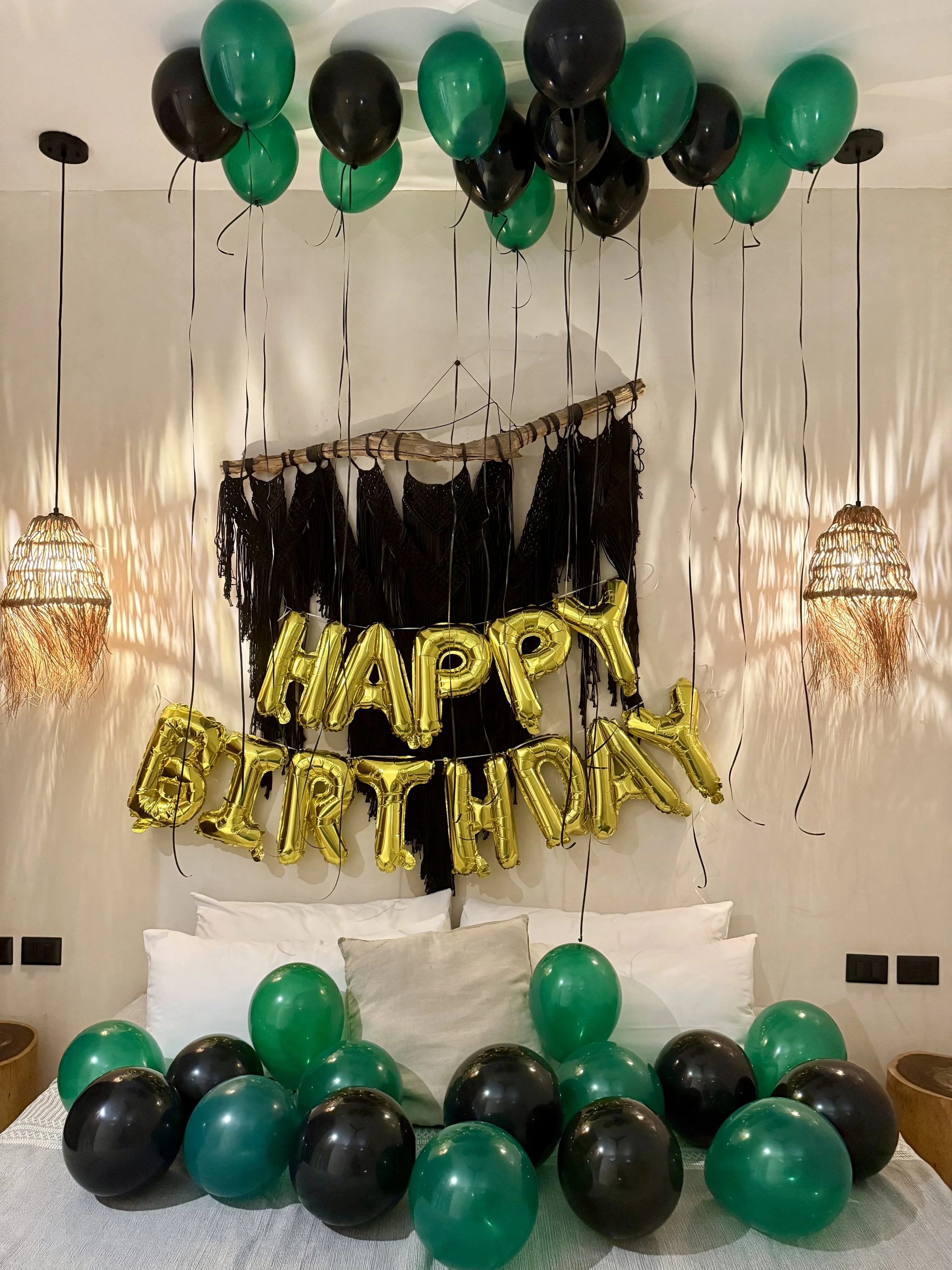 Room decorated for a birthday celebration with black and green balloons, a 'Happy Birthday' gold foil balloon banner, and hanging lights.
